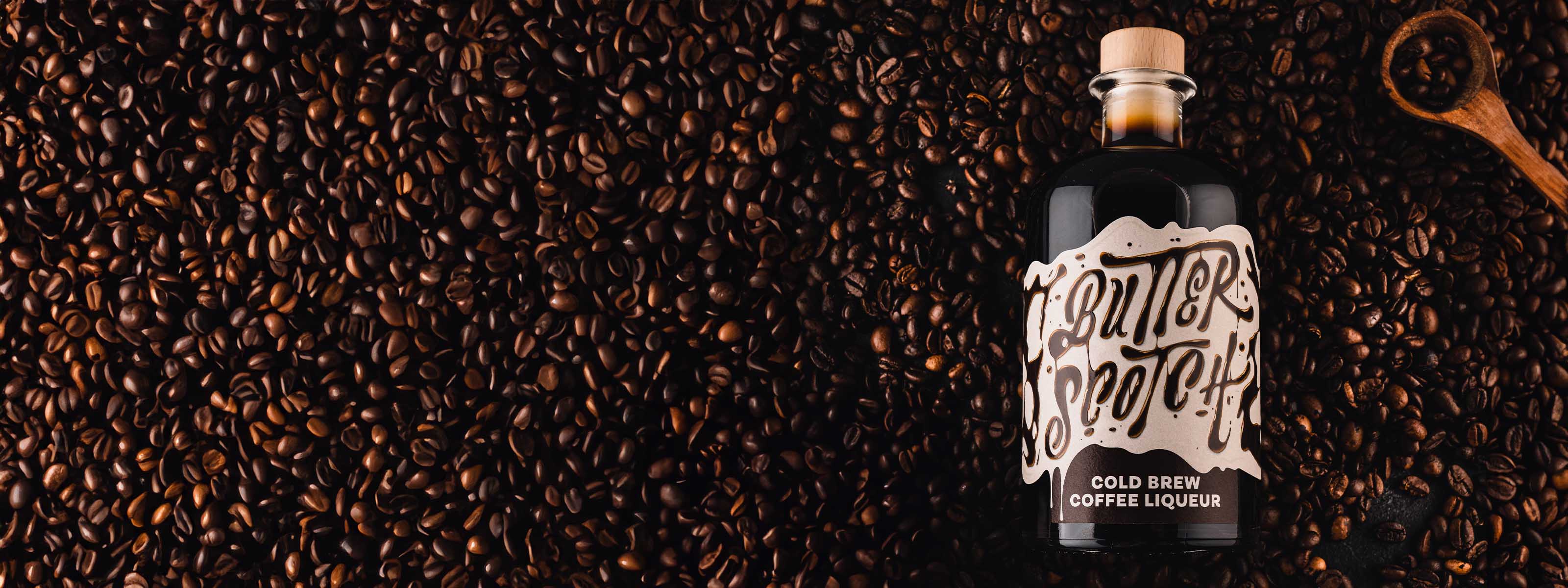 Butterscotch Cold Brew Coffee Likör Flasche liegend in dunklen Kaffeebohnen, kleiner Portionierungslöffel daneben, fotografiert von oben