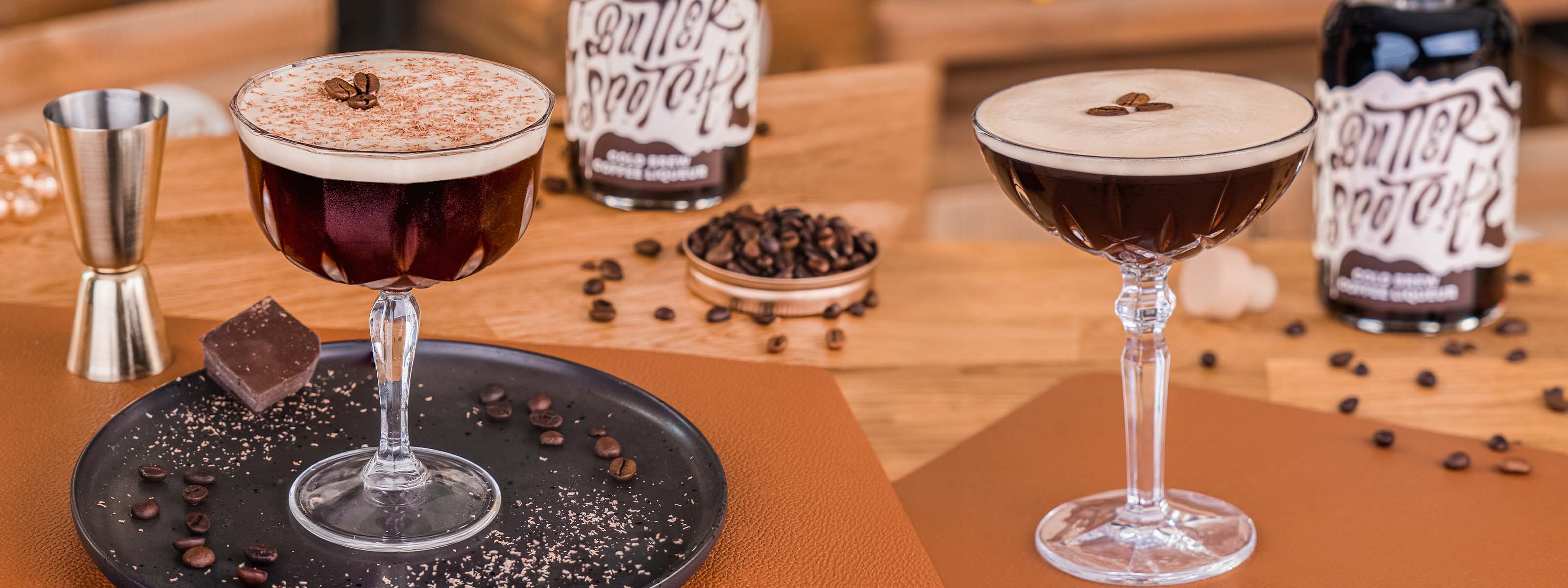 Butterscotch Espresso Martini Drink, Espresso Martini im geschliffenen Stielglas, mit Schaumkrone, dekoriert mit 3 Kaffeebohnen, auf hellem Untergrund, Kaffeebohnen, Messbecher und Kaffeebohnen im Hintergrund