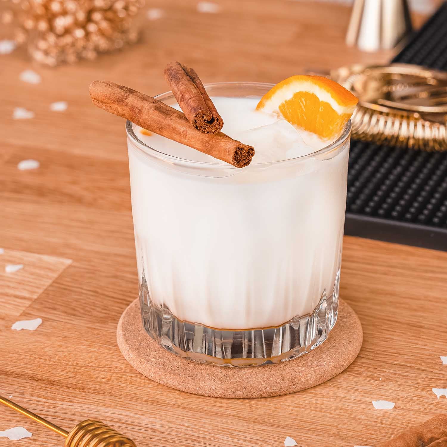 Honig, Milch & Whiskey Drink, weißer Cocktail im geschliffenen Glas im Vordergrund, dekoriert mit Zimtstangen und Orangenscheibe, vor hellem Bar-Hintegrund