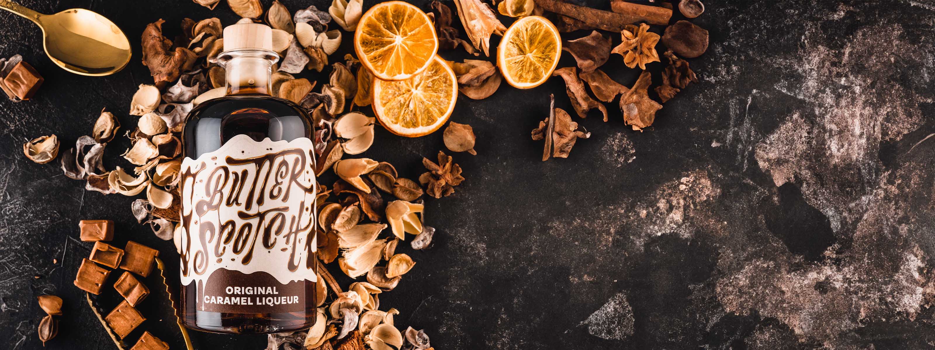 Flasche Original Caramel Likör liegend zwischen Karamellbonbons, Gewürzen und getrockneten Orangenscheiben auf dunklem Untergrund, von oben fotografiert.