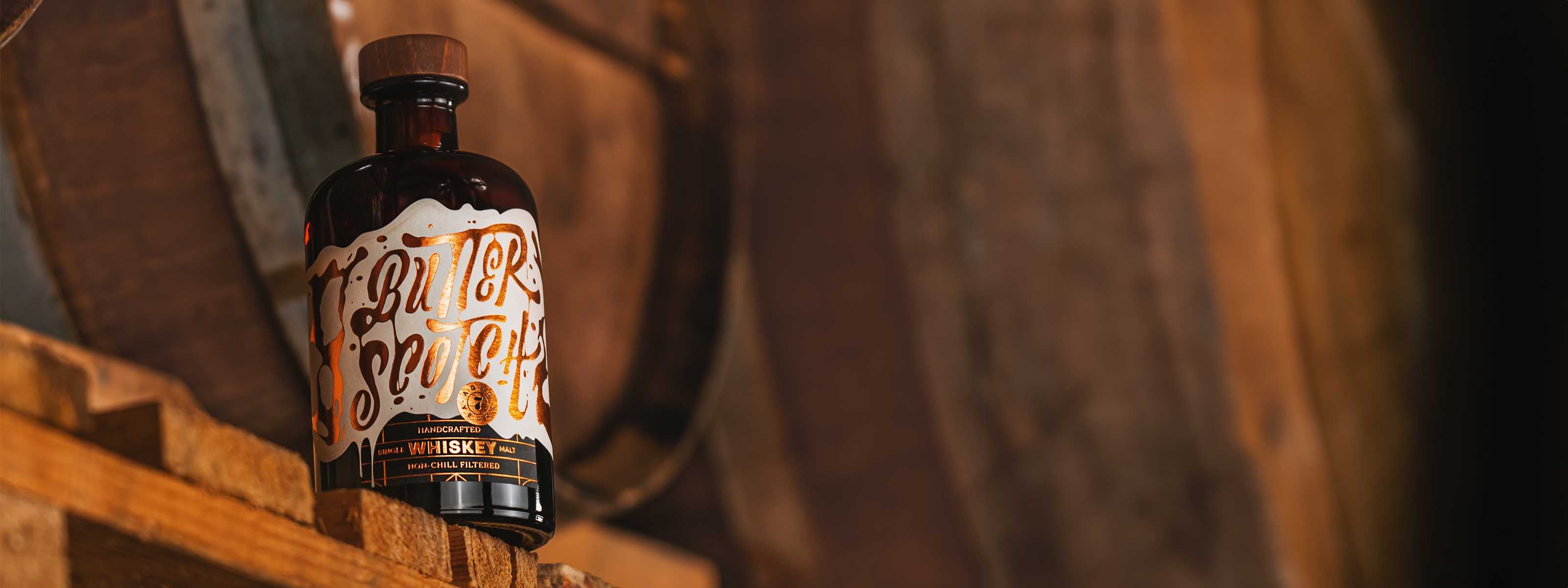 Butterscotch Single Malt Whiskey, bronze glänzendes Etikett, Flasche mit Holzkork, vor großen Whiskey-Fässern im Hintergrund