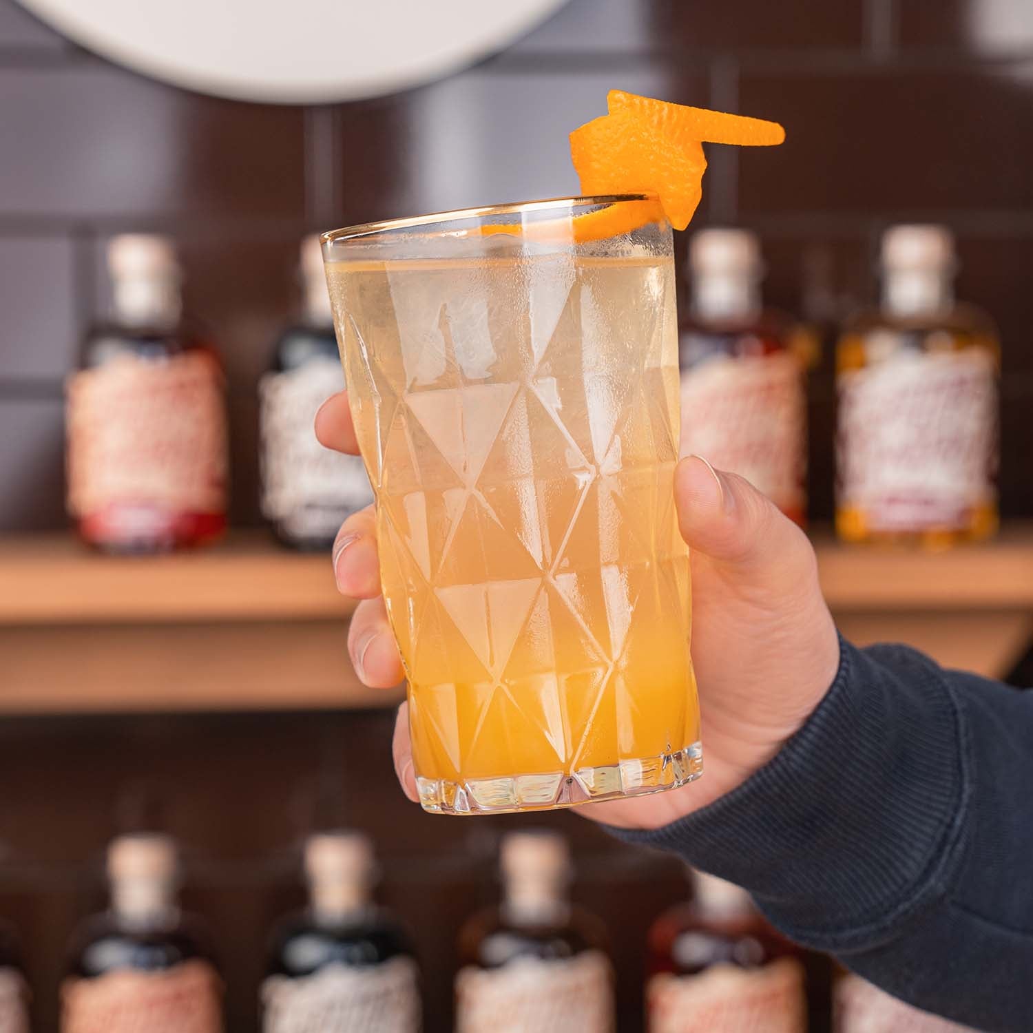 Butterscotch Bratapfel Bonnies Love Cocktail, Longdrinkglas gefüllt mit Bratapfel Likör, Orangensaft und Chmpagner. Dekoriert mit einer Orangenzeste, vor Butterscotch Flaschen im Hintergrund