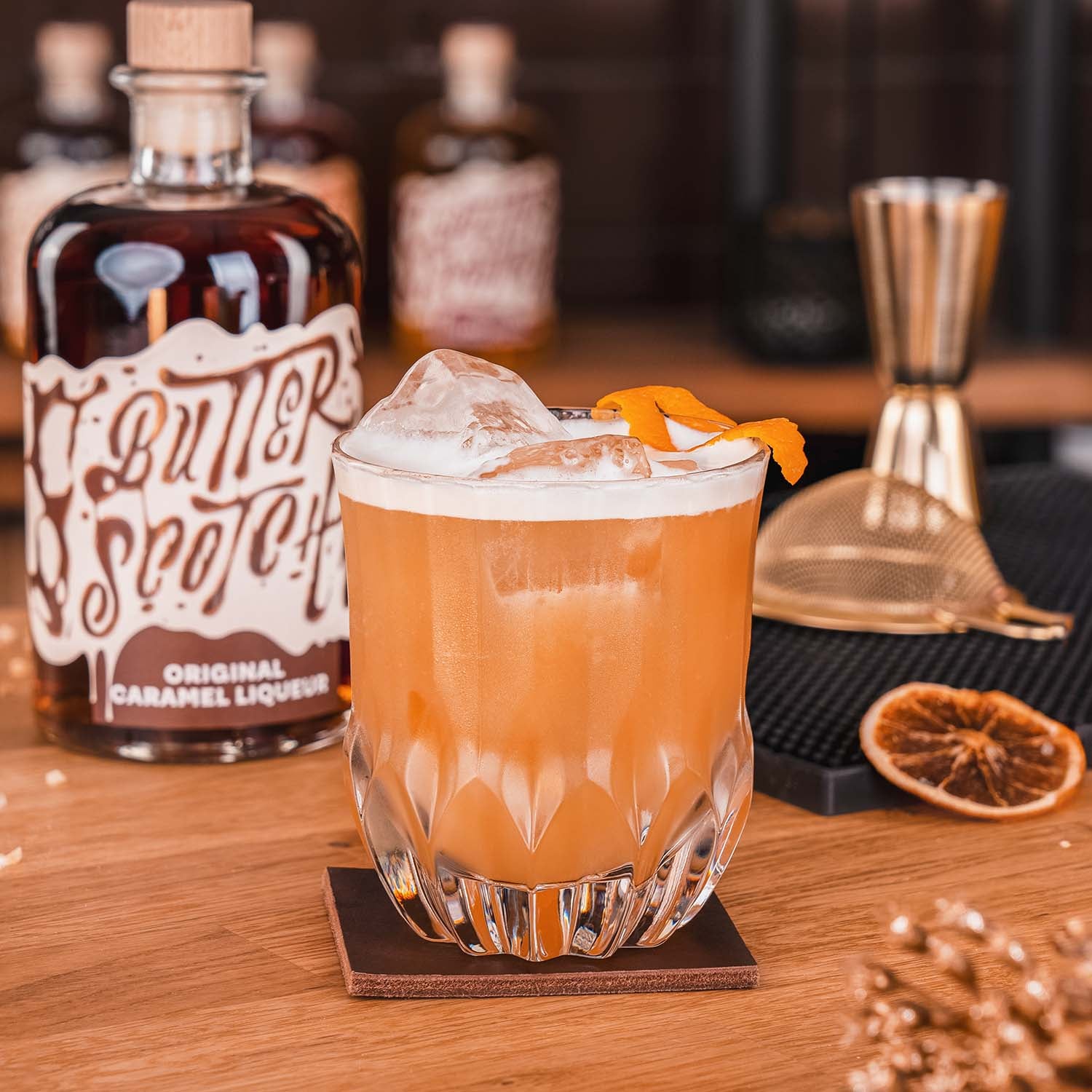 Butterscotch original Caramel Liqueur Flasche unscharf im Mittelgrund, Caramel Sour Cocktail im Vordergrund, Orangener Drink in einem mit Eis gefüllten Glas, dekoriert mit einer Orangenzeste