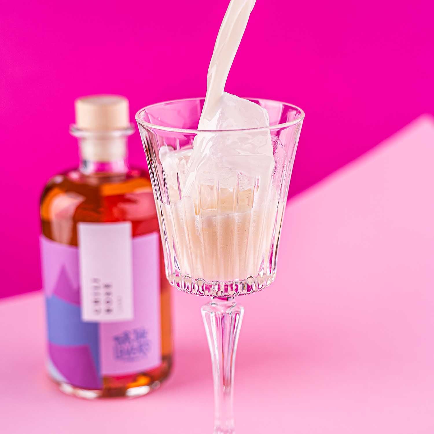 For the Lovers Chilli Rose Sour Cocktail wird in eine Glas auf Eis eingegossen, Flasche Chilli Rose Likör im Mittelgrund auf pink / rosanem Untergrund