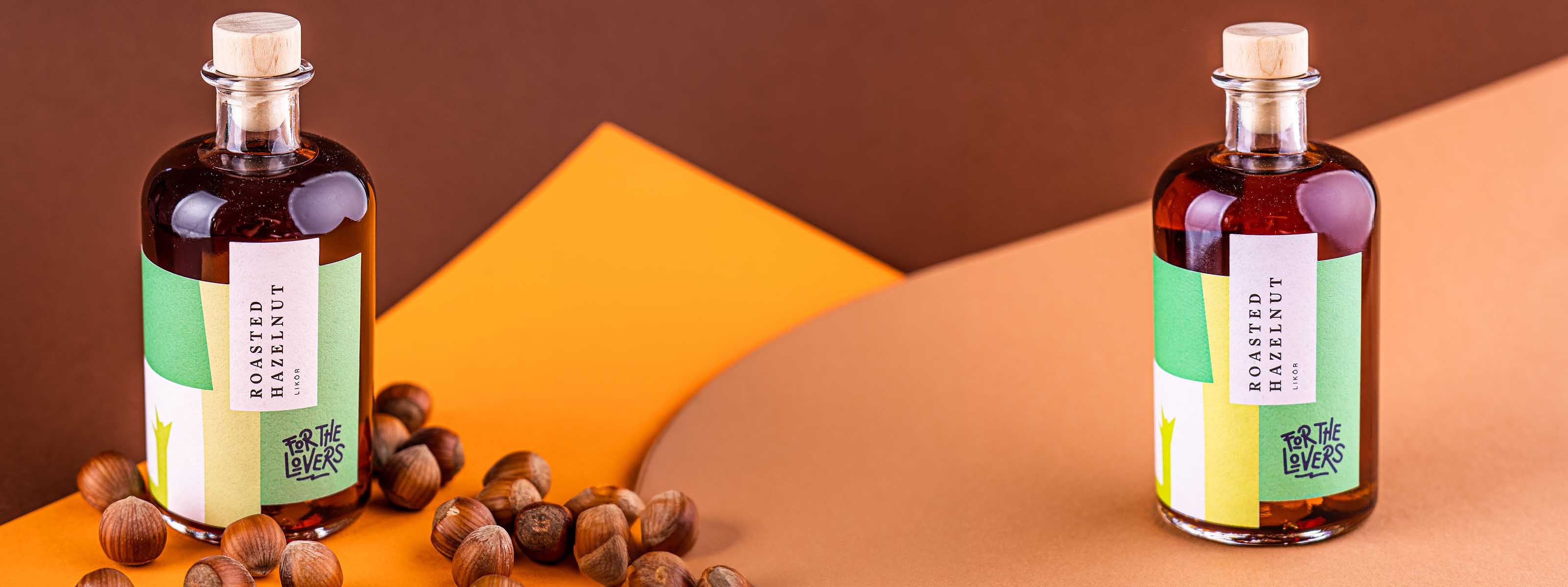 For the Lovers Hazelnutt Likör Flaschen im Vordergrund zwischen Haselnüssen auf gelb / braunem Untergrund