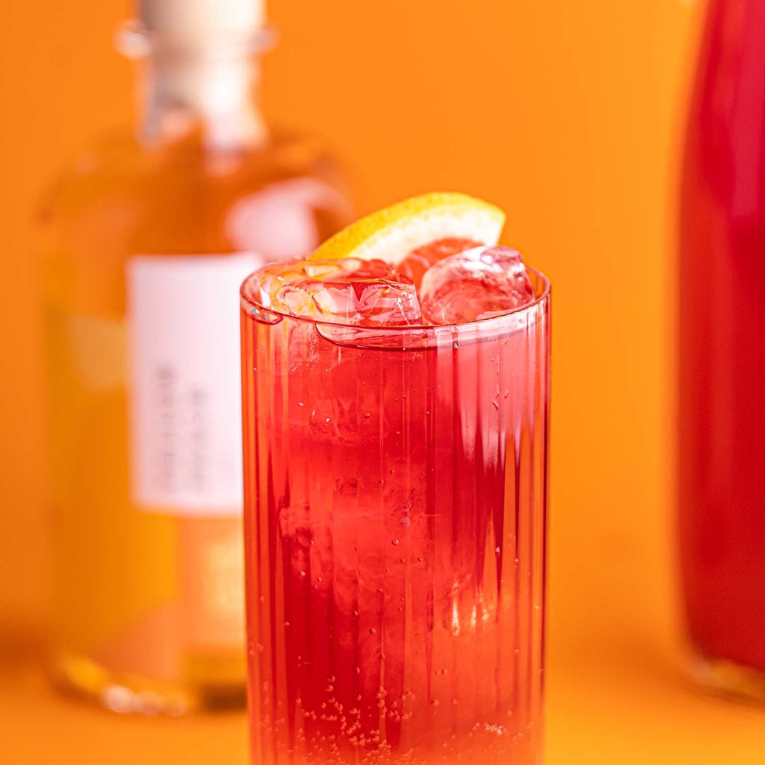 Orangener Longdrink in geriffeltem Glas im Vordergrund, dekoriert mit frischer Grapefruit-Scheibe, Flasche Yellow Peach Likör angeschnitten im Hintergrund auf orangenem Untergrund