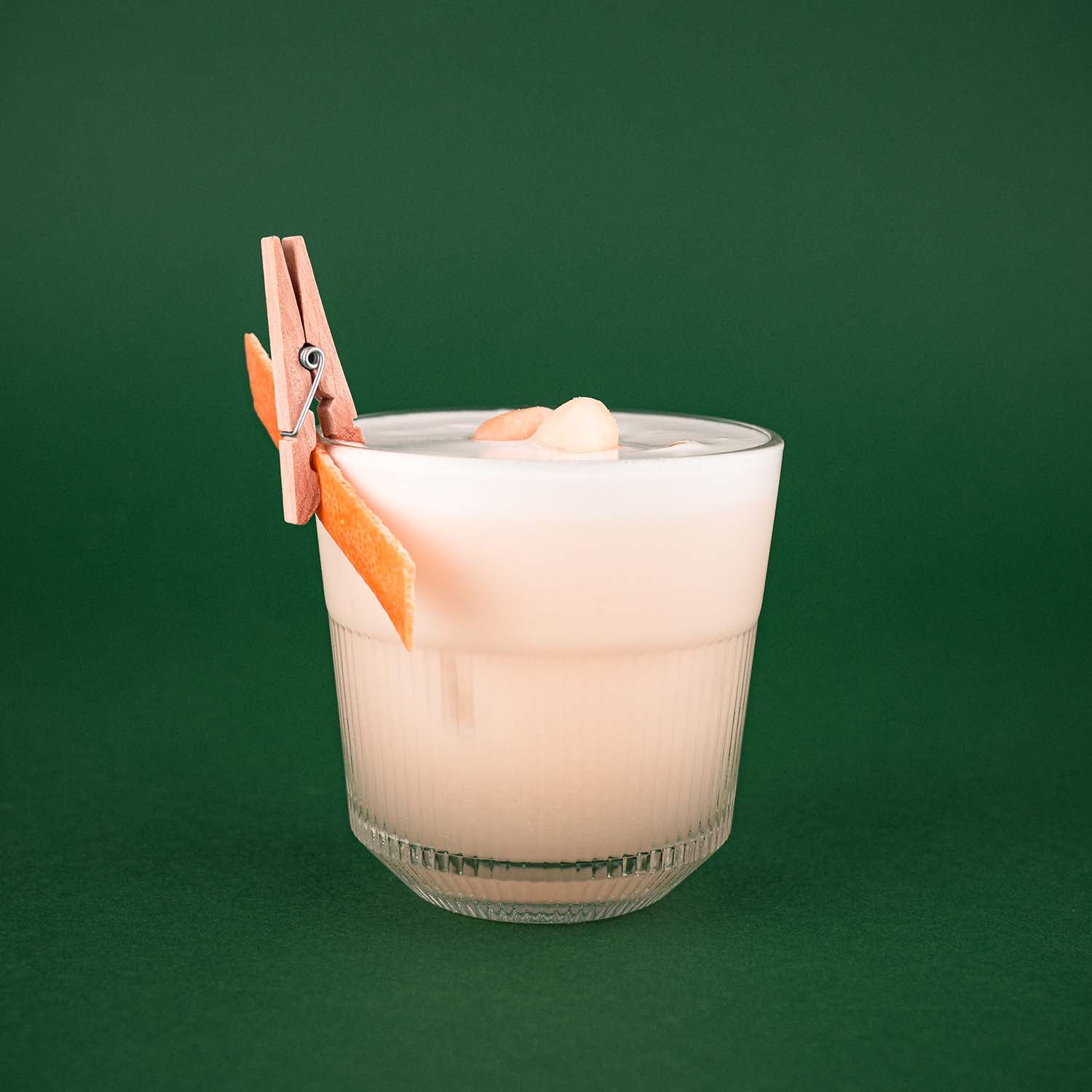 Hazellujah Macadamia Sour Cocktail, Weißer Cocktail mit Schaumkrone, dekoriert mit Macadamia-Nüssen und einer Orangenzeste, vor dunkelgrünem Hintergrund