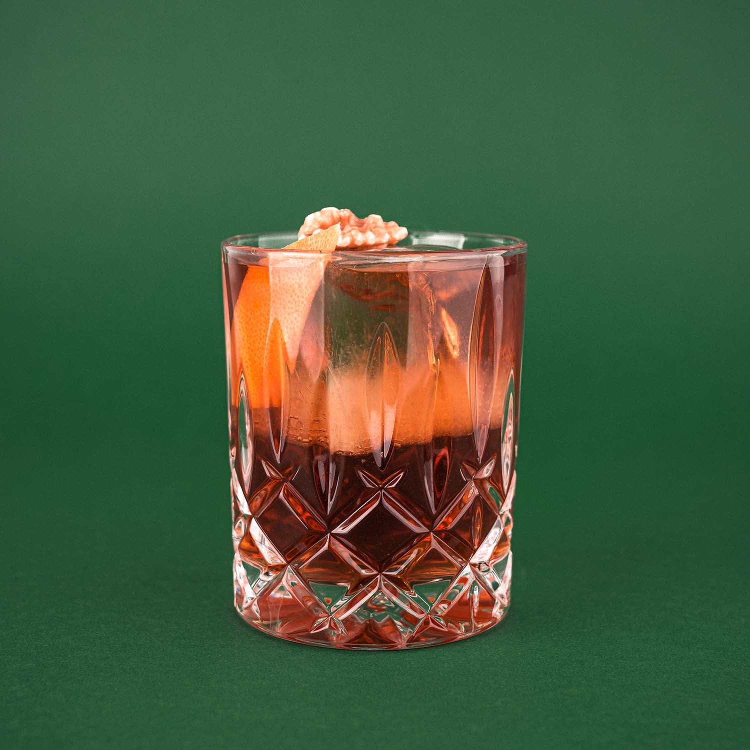 Hazellujah Walnut Old Fashioned Cocktail, Dunkler Drink im geschliffenen Glas, dekoriert mit Orangenzeste und Walnuss, vor dunkelgrünem Hintergrund