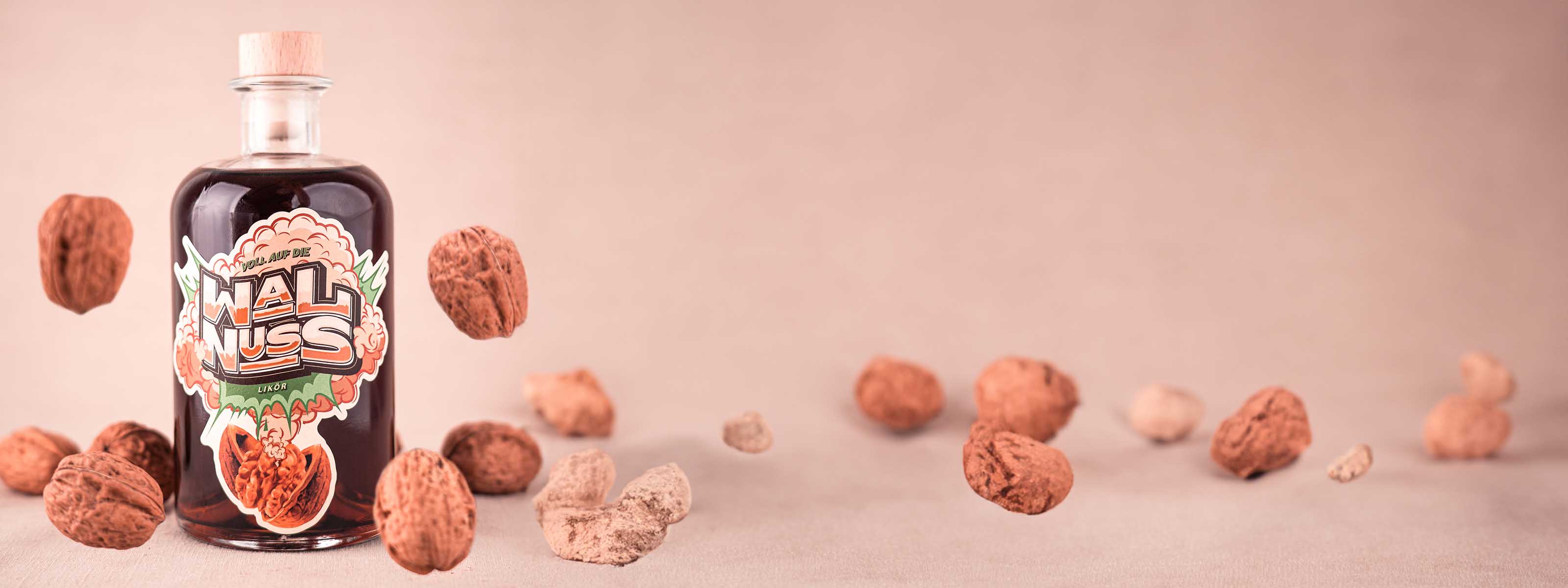 Hazellujah Walnuss Likör, Flasche Likör im Vordergrund zwischen fallenden Walnüssen auf hellem Untergrund
