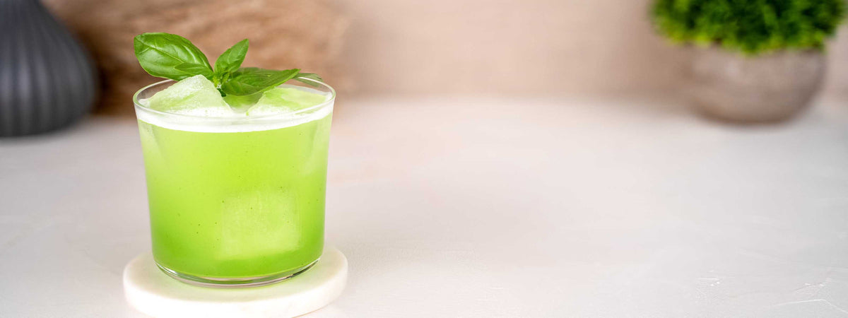 Momotaro Gin Basil Smash Cocktail, grüner Cocktail im schlichten Glas mit Eiswürfeln, dekoriert it frischer Minze, Trockenblumen und Pflanze im Hintergrund auf hellem Untergrund