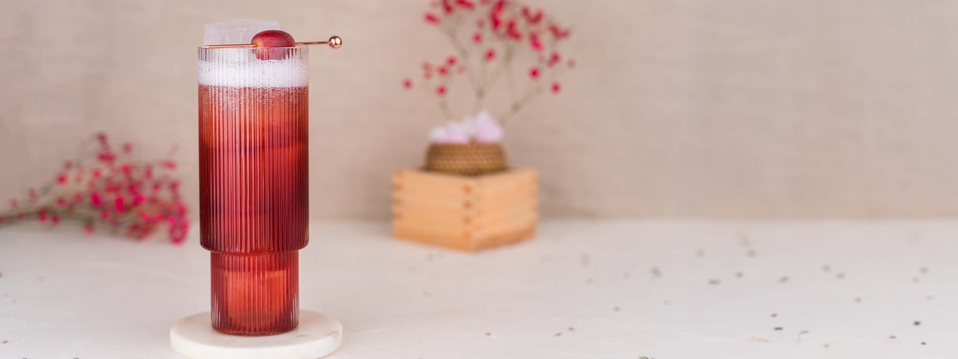 Momotaro Akainu Cocktail mit Traubensaft und Tonic water, Longdrinkglas mit rotem Inhalt, garniert mit einer Traube, auf hellem Untergrund mit Blüten im Hintergrund