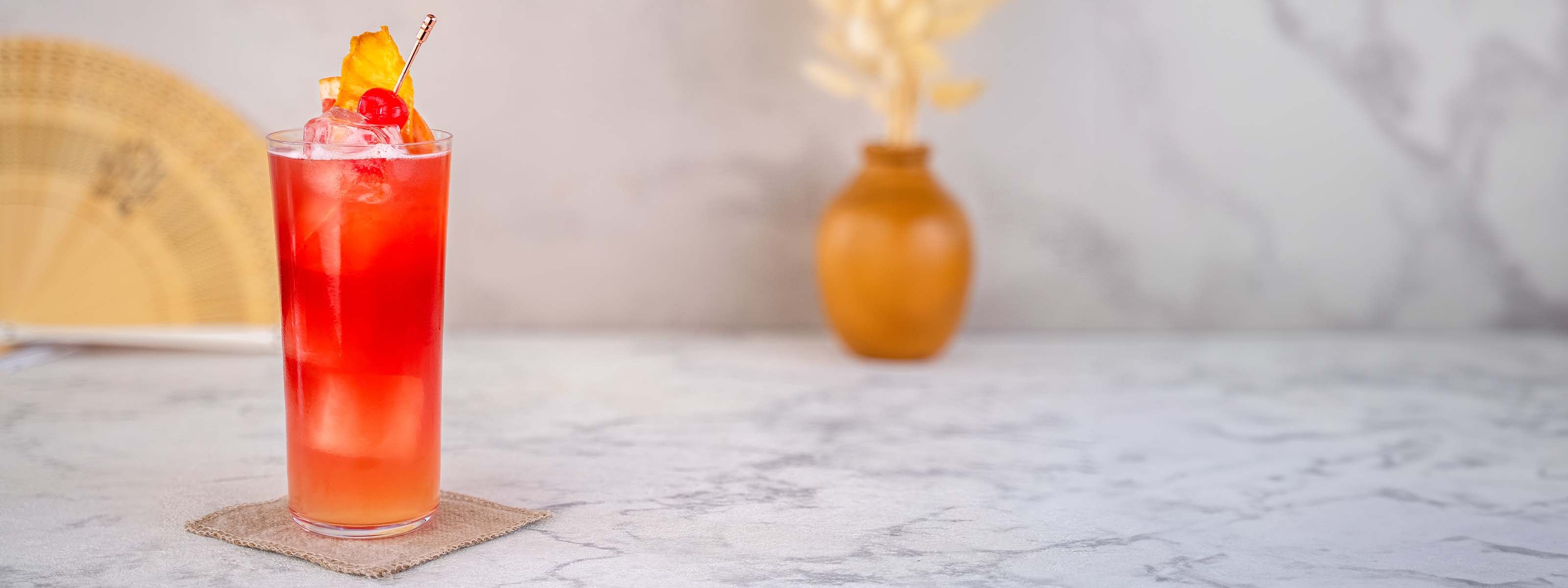 Momotaro Gin Tokio Sling Cocktail, orangefarbener Cocktail im Longdrinkglas mit Eiswürfeln, dekoriert mit Orangenscheibe und Cocktail-Kirsche, Vase und japanischer Fächer als Dekorationselemente im Hintergrund, auf hellem Marmor-Untergrund