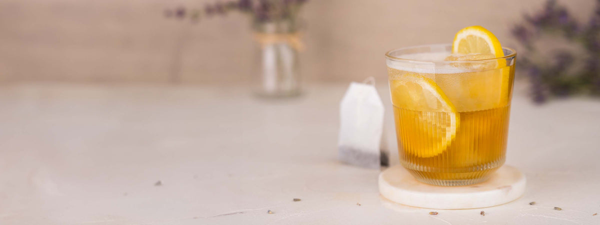 Momotaro Gin & Tea Cocktail, Drink mit Momotaro Ginzero und Tee, gelber Drink mit Zitronenscheiben garniert, Teebeutel und Lavendelblüten im Hintergrund auf hellem Untergrund