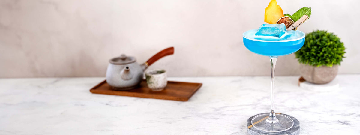 Momotaro Blue Ocean Cocktail, blauer Drink in einer Coupette, mit großen Eiswürfel in der Mitte des Glases, dekoriert mit frischer Minze und getrockneter Limettenscheibe, traditionell japanisches Tee-Set im Hintergrund auf hellem Untergrund