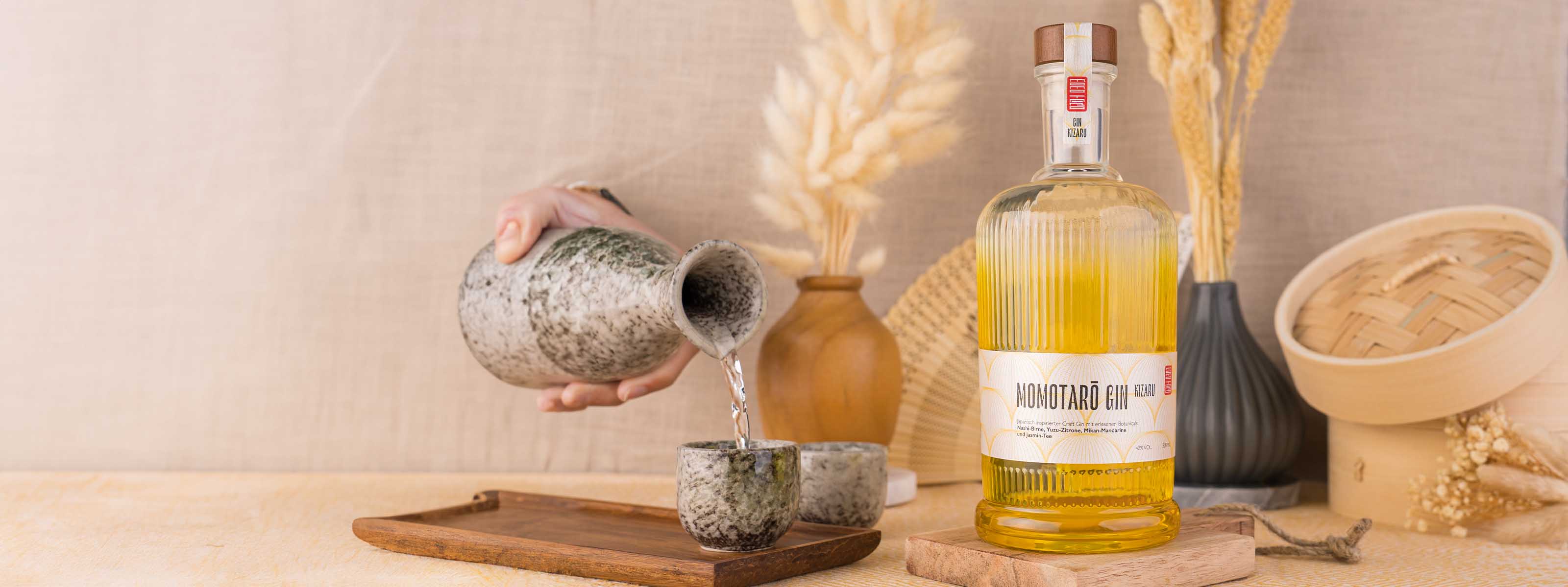 gelbe Momotaro Kizaru Flasche im Vordergrund, Sake wird in Steinbecher eingegossen, Vase mit Trockenblumen im Hintergrund, auf hellem Jute-Stoff