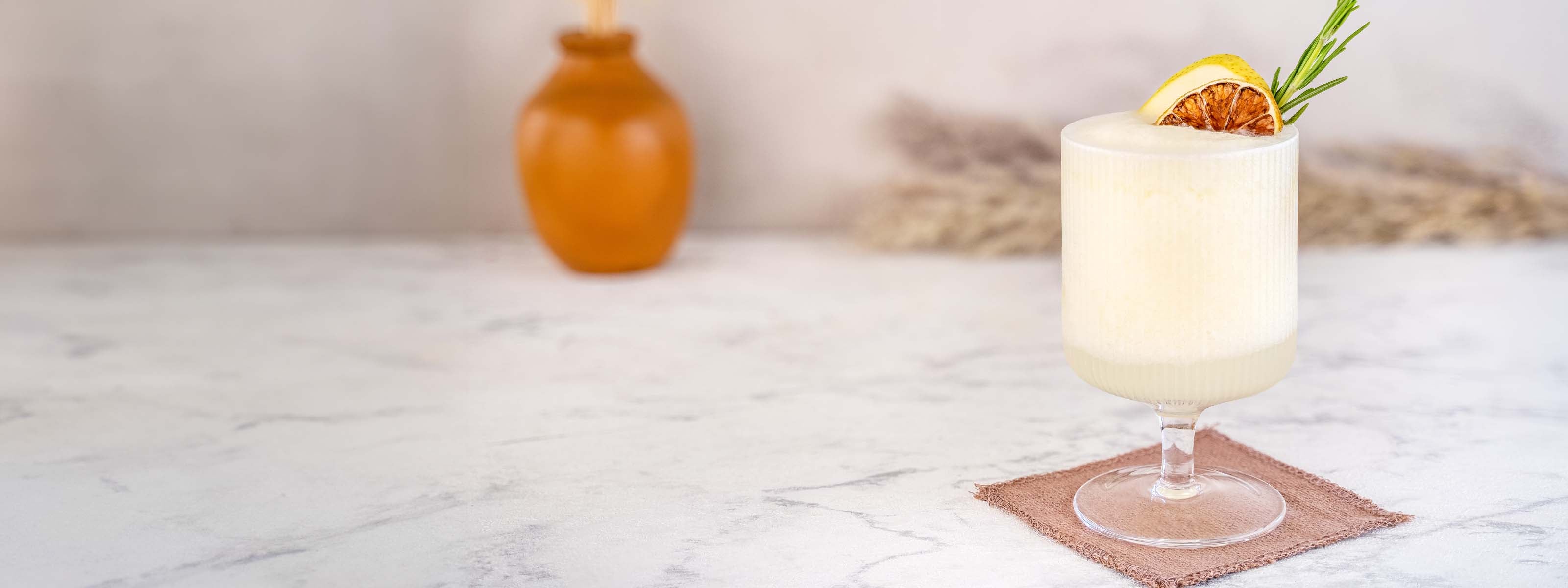 Momotaro Kizaru Slushy Drink, gelber Drink mit Eis und pürierter Birne, dekoriert it Birne, getrockneter Limette und Thymianzweig vor hellem Hintergrund