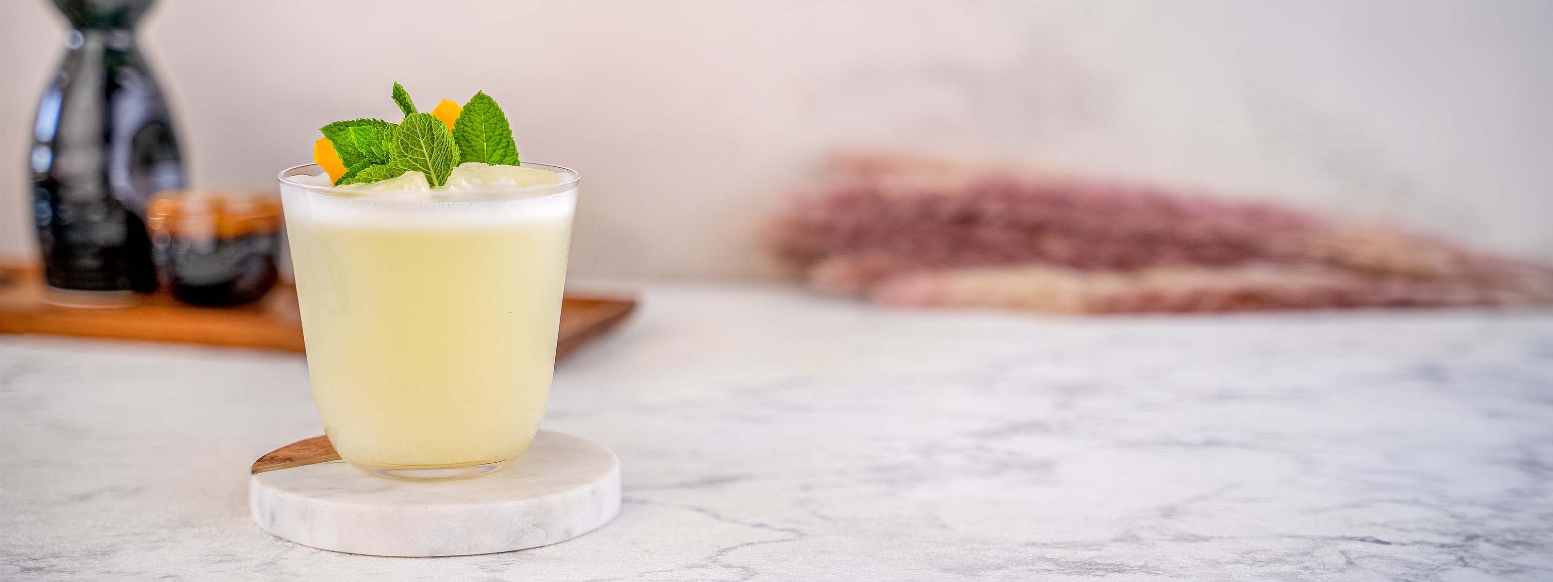 Momotaro Kizaru Smash Drink, Gelber Drink in schlichtem Glas auf einer Marmorplatte, dekoriert mit frischer Minze und einer Orangenzeste, japanisches Sake Set im Hintergrund auf hellem Untergrund