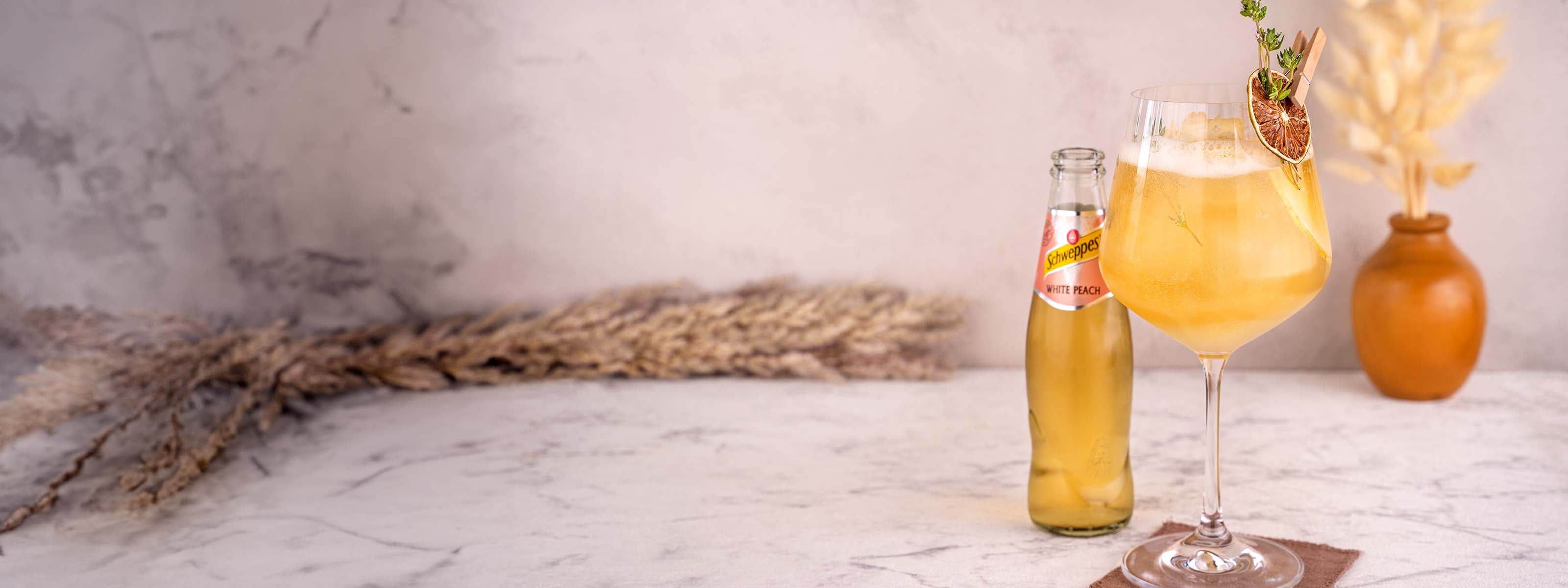 Momotaro Kizaru Spritz Cocktail, Großes Glas mit gelbem Drink, garniert mit Limettenscheibe und Thymian, Schweppes White Peach Limonade im Mittelgrund, Vase und Trockenblumen als Dekorationselemente im Hintergrund