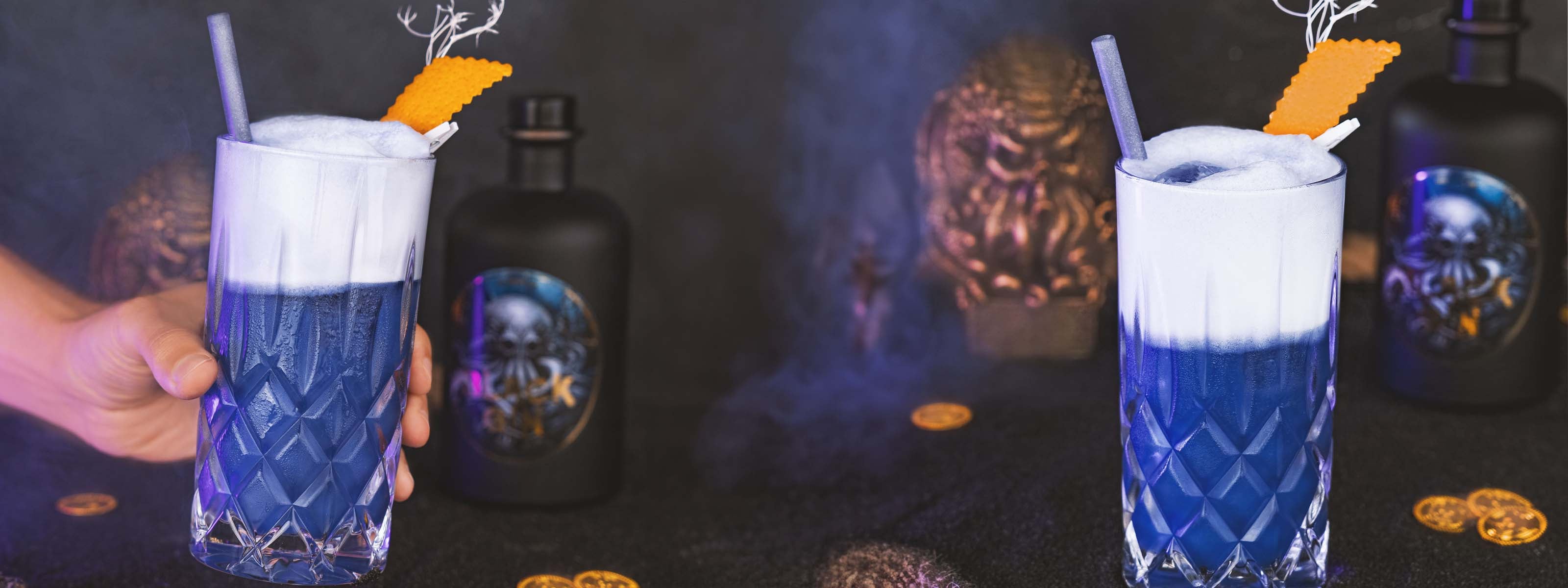 Sepia Ink Gin Fizz Cocktail, dunkelblauer Cocktail mit weißer Schaumkrone und Orangenschale vor dunklem Hintergrund mit nautischen Elementen