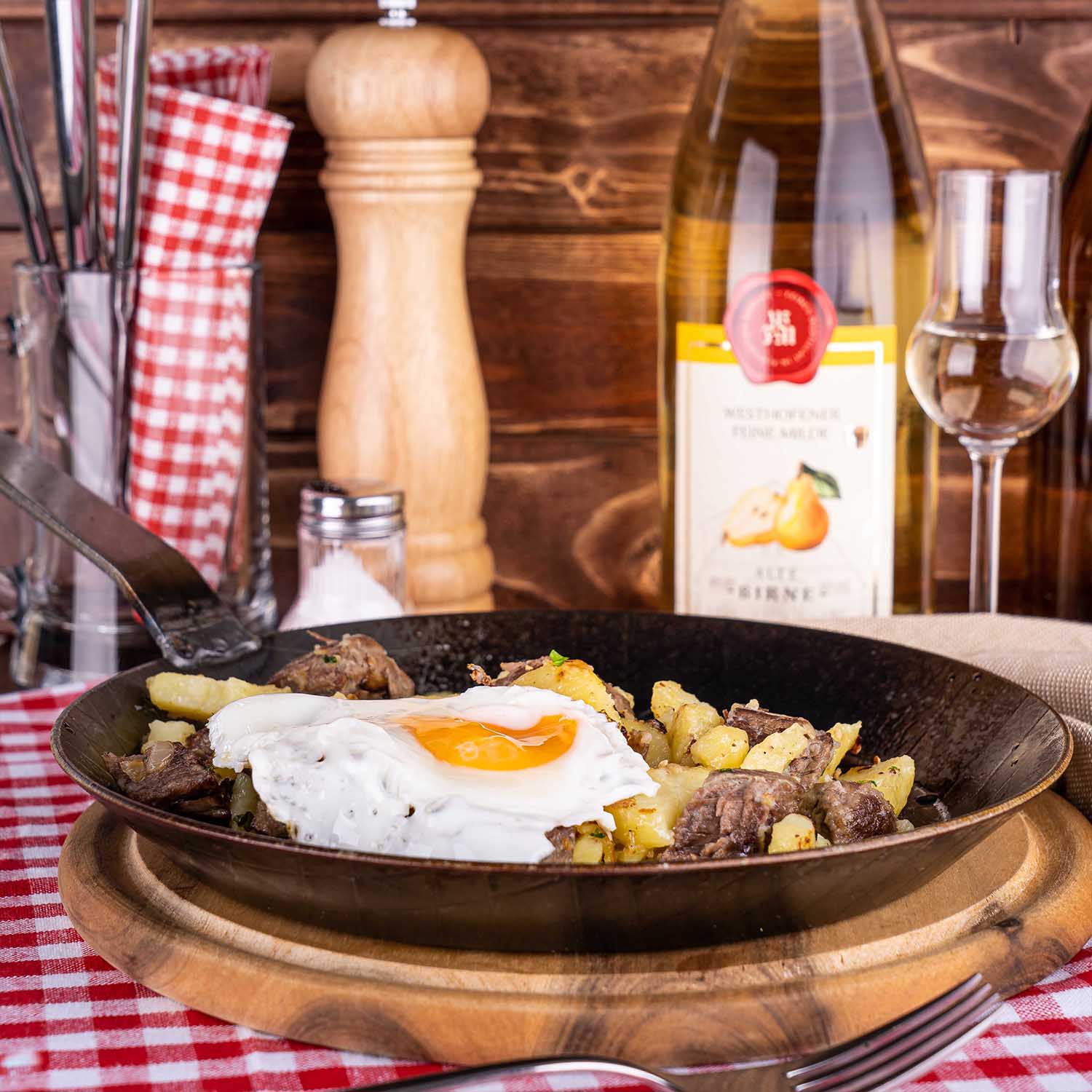 gusseiserne Pfanne mit Kartoffeln, Fleisch und Spiegelei im Vordergrund, Westhofener Feine Milde Birnenbrand im Hintegrund, auf rot weiß karierter Tischdecke, vor Holzlatten im Hintergrund