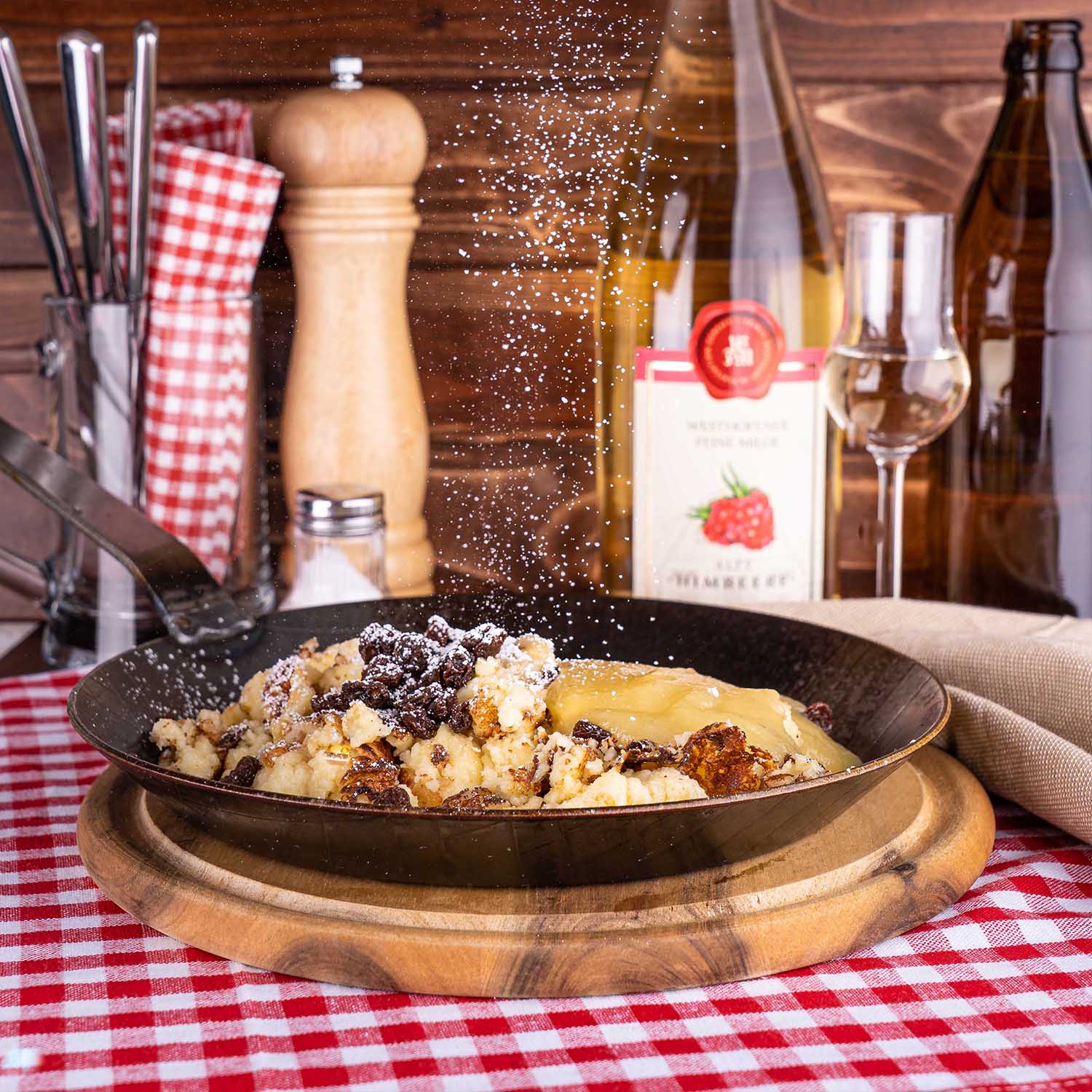gusseiserne Pfanne mit Kaiserschmarren im Vordergrund, Puderzucker fällt von oben auf die Pfanne herunter, Westhofener Feine Milde Himbeerbrand mit Nosing-Glas im Hintergrund, auf rot/weiß karierter Tischdecke vor dunklen Holzlatten im Hintergrund