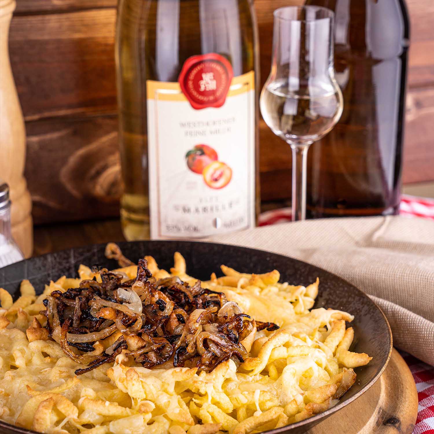 Käsespätzle mit Schmelzzwiebeln in gusseiserner Pfanne im Vordergrund, Westhofener Feine Milde Alte Marille mit Nosing-Glas im Hintergrund, auf karierter Tischdecke vor Holzlatten im Hintergrund