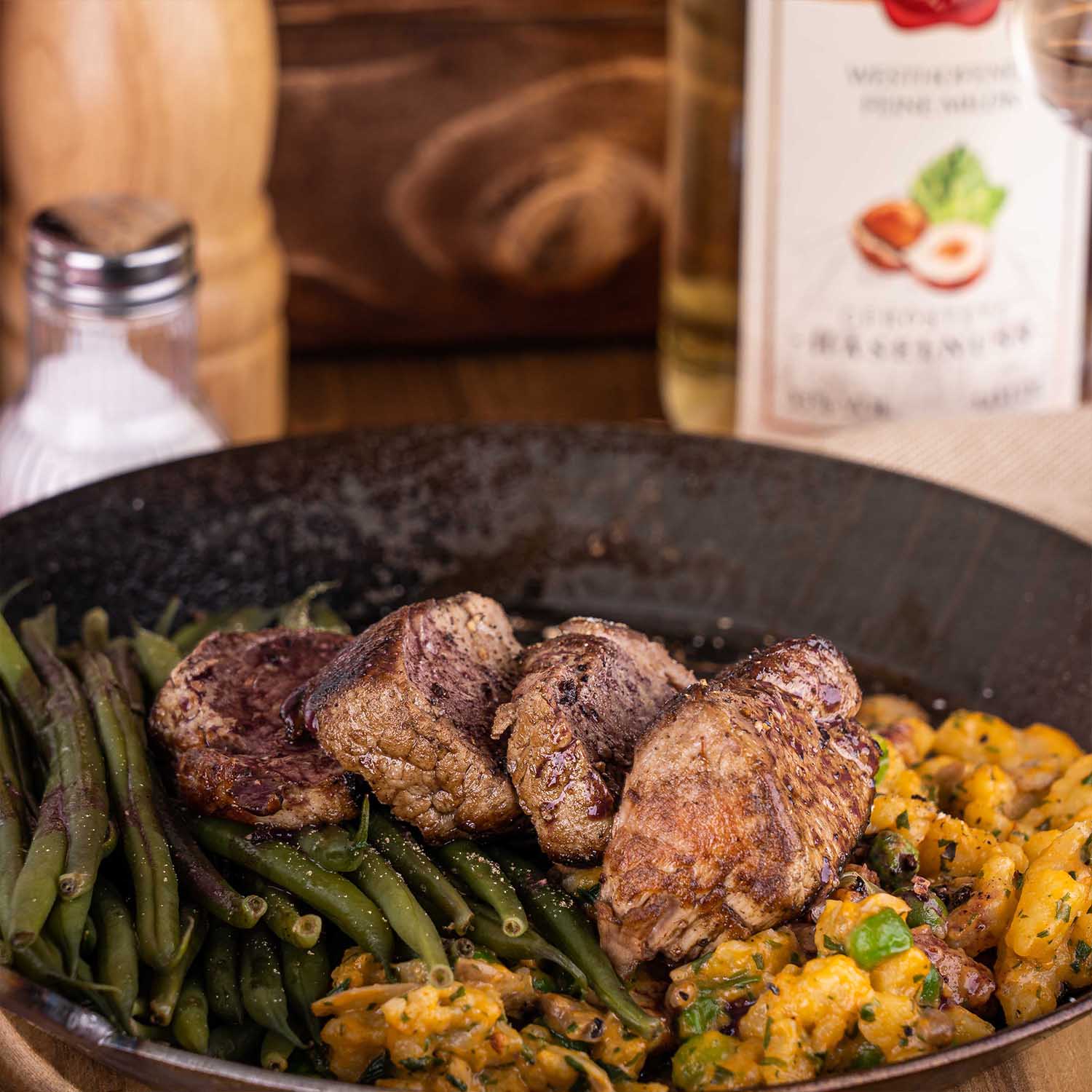 Gusseiserne Pfanne mit gebratenem Fleisch, Bohnen und Kartoffeln im Vordergrund, Westhofener Feine Milde geröstete Haselnuss angeschnitten im Hintergrund