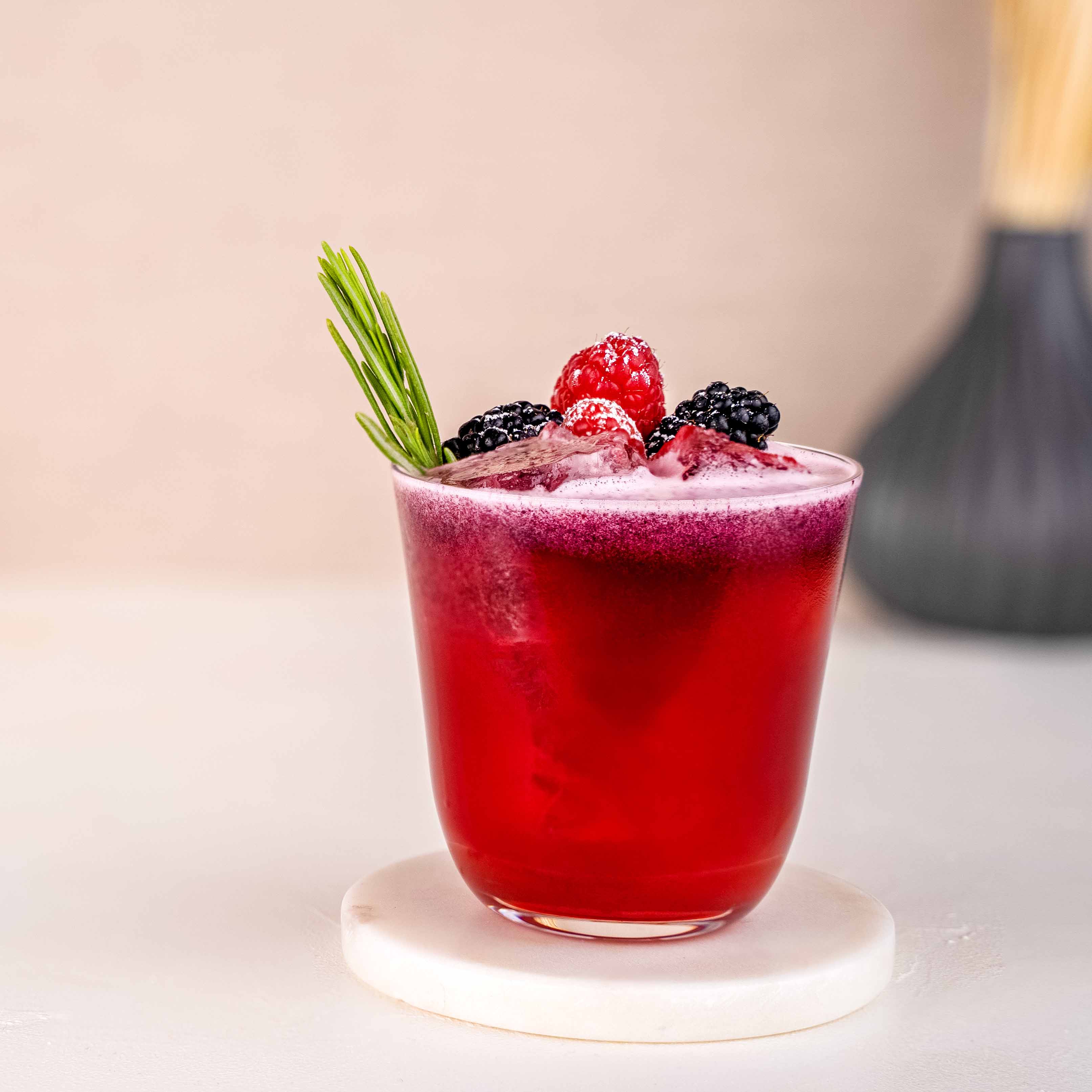 Momotaro Akainu Bramble Cocktail, roter Drink mit frischen Beeren und Thymian garniert, auf weißem Untergrund, Vase und helle Dekorationselemente im Hintergrund