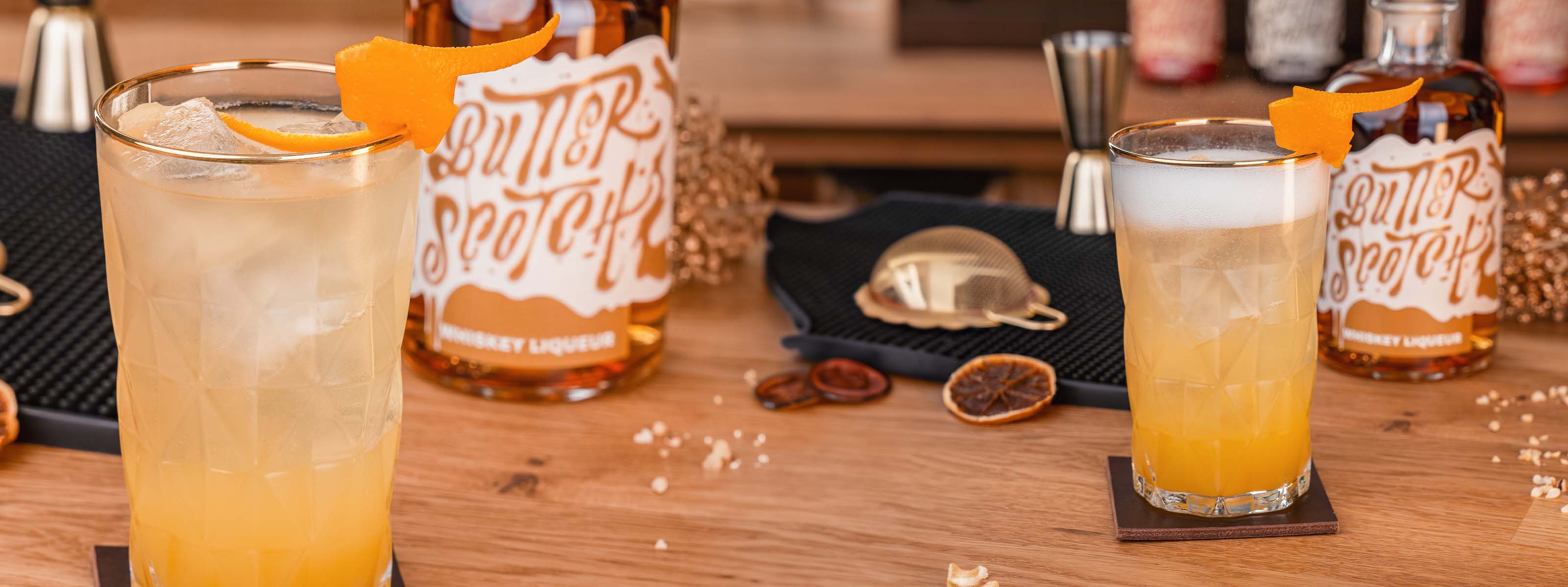 Butterscotch Bonnies Love Drink mit viel Eis angeschnitten im Vordergrund, dekoriert mit Orangenzeste, Butterscotch Whiskey Likör Flasche, Barmatte mit Barbesteck im Hintergrund