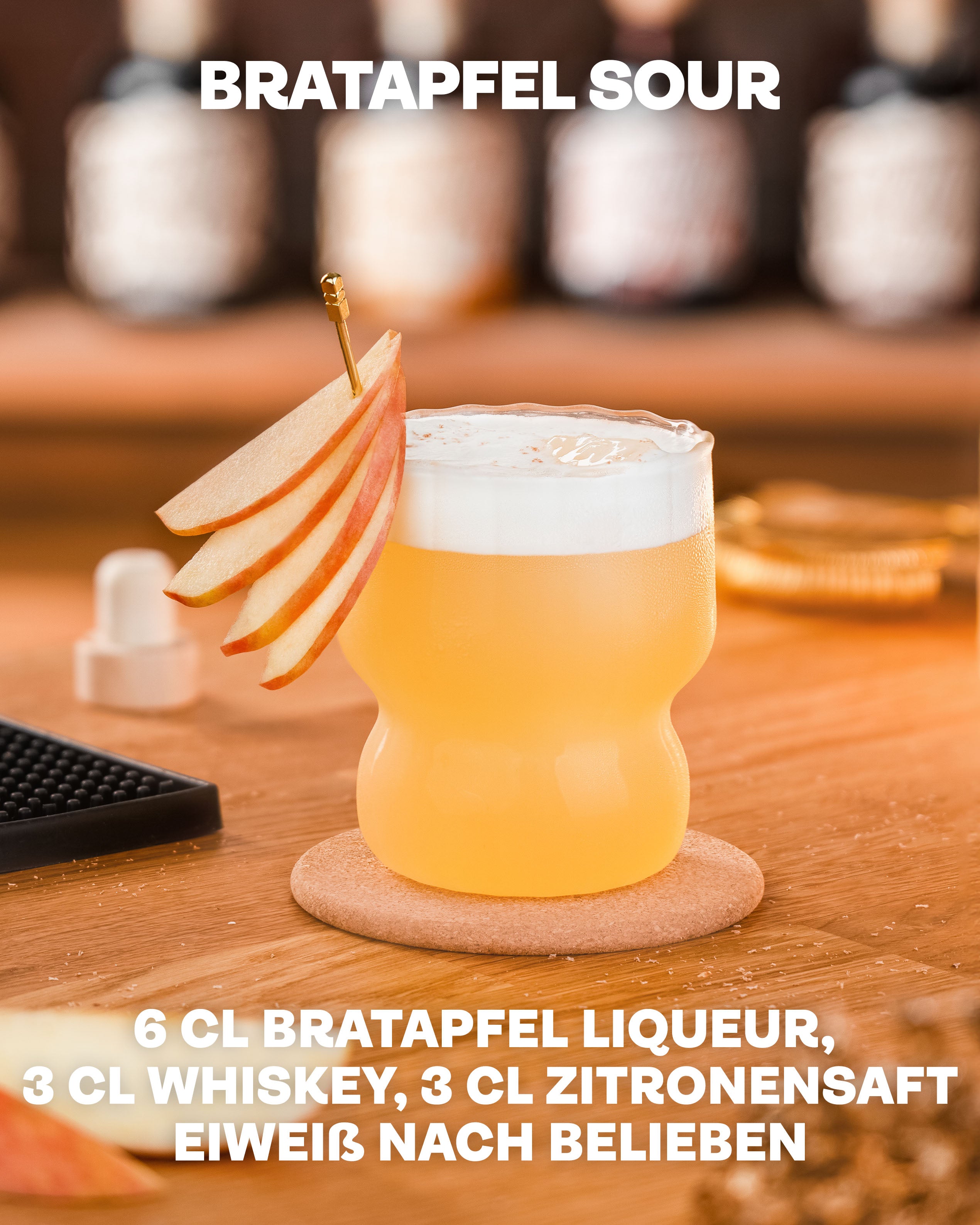 Butterscotch Bratapfel Sour Drink, Bratapfel Likör mit Whiskey und Zitronensaft, Drink auf Eis im Vordergrund, dekoriert mit Apfelscheiben auf hellem Untergrund, Bar unscharf im Hintergrund