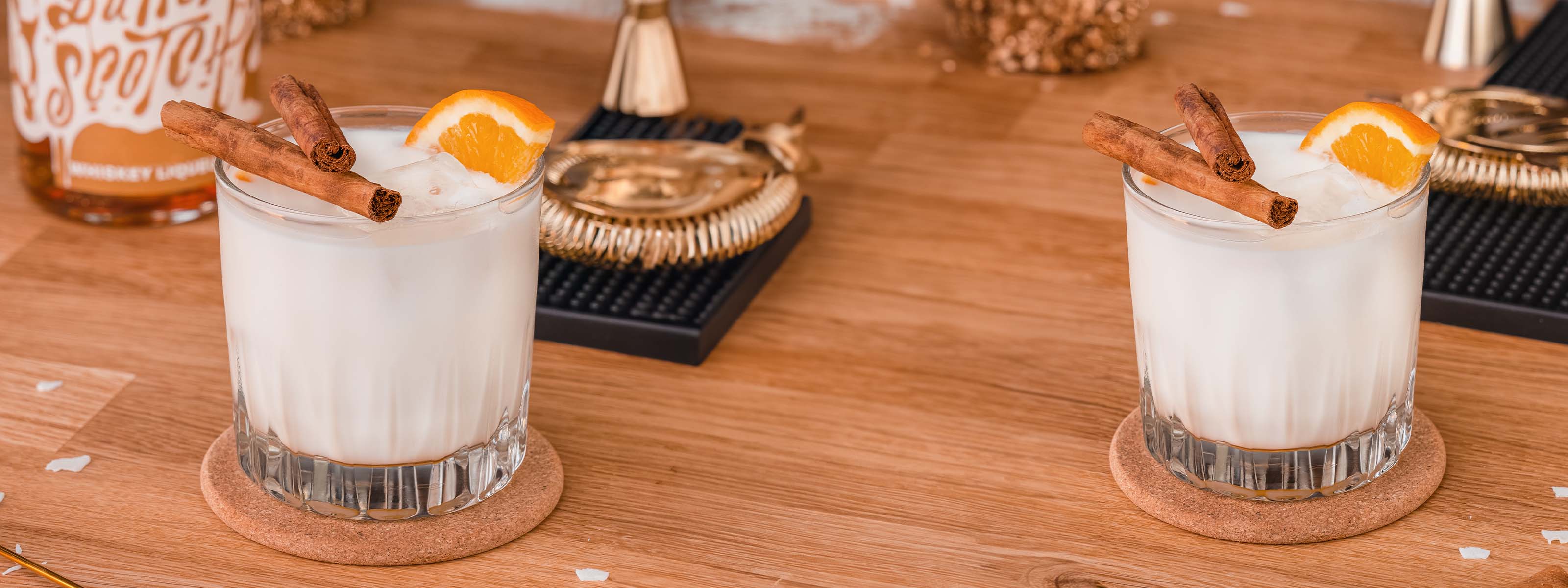 Honey & Milk Cocktail, Milch Cocktail im geschliffenen Glas mit Eis, dekoriert mit Zimtstangen und Orangenscheibe, Flasche Whiskey Likör, Barmatte & Barbesteck unscharf im Hintergrund