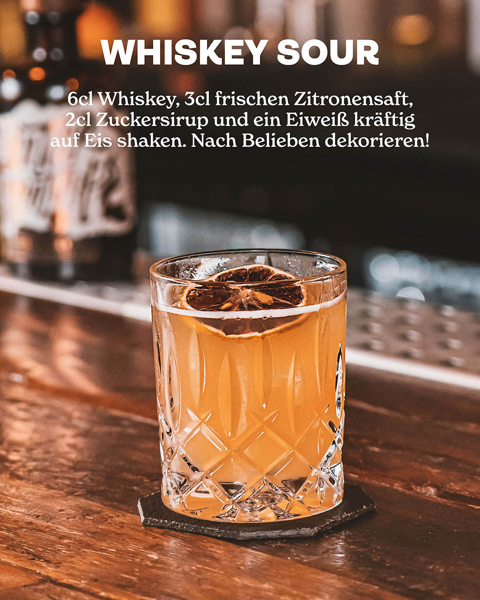 Butterscotch Whiskey Sour, Whiskey, Zitronensaft und Zuckersirup auf Eis, Whiskey Sour in geschliffenem Glas auf dunklem Holztresen, dekoriert mit getrockneter Limette, Whiskeyflasche unscharf im Hintergrund