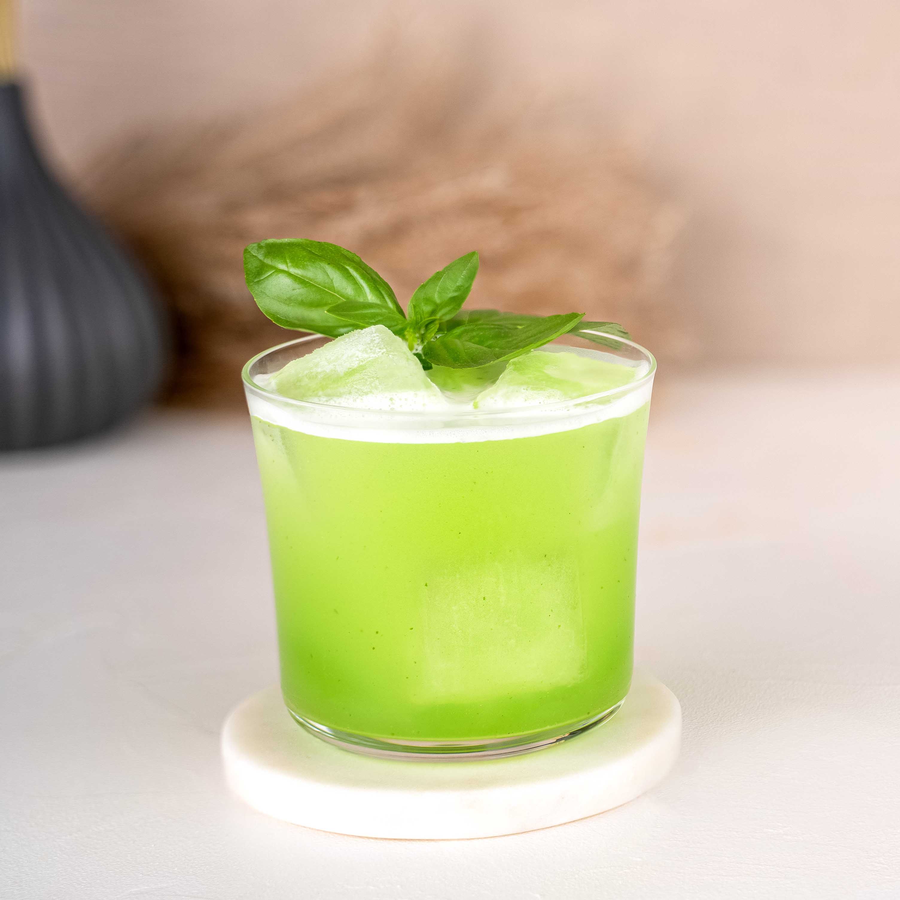 Momotaro Basil Smash Cocktail, grüner Cocktail im schlichten Glas, dekoriert mit frischen Basilikumblättern, auf hellem Untergrund, Trockenblumen als Dekoration im Hintergrund