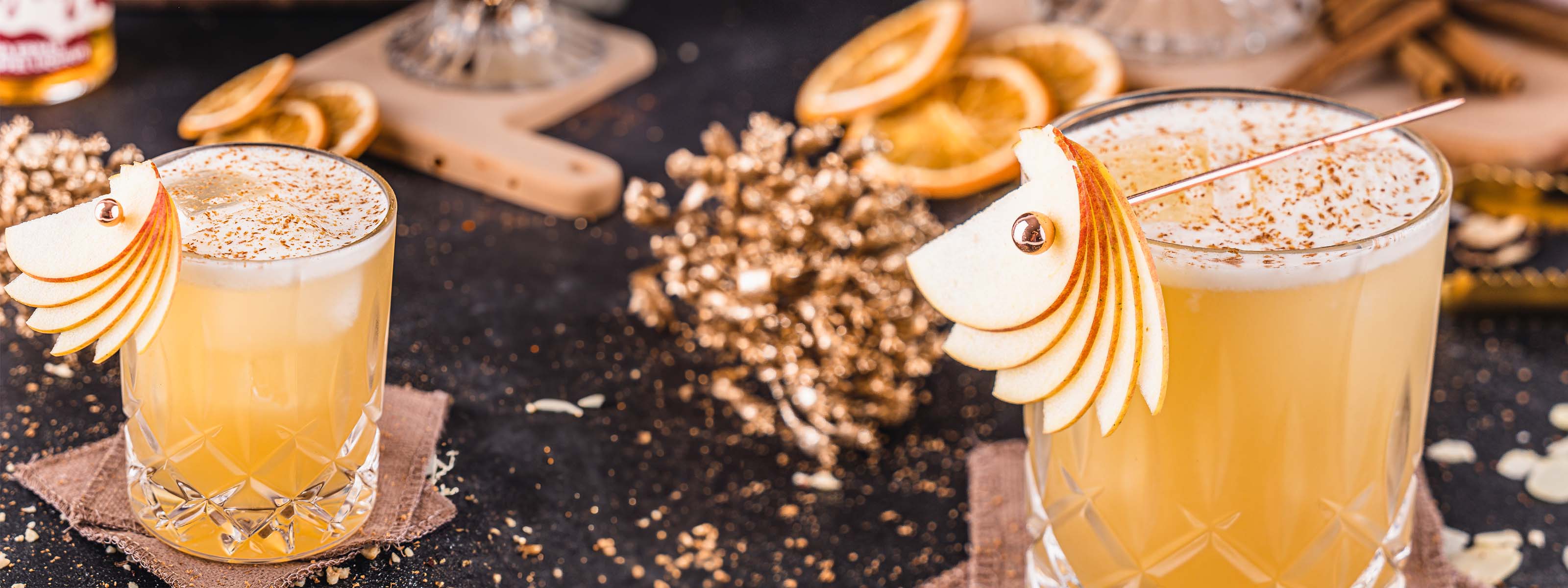 Butterscotch Bratapfel Sour Cocktail, Gelber Drink im geschliffenen Glas auf Eiswürfeln, mit Schaumkrone, dekoriert mit Apfelscheiben und Schokoladenraspeln, auf dunklem Untergrund 