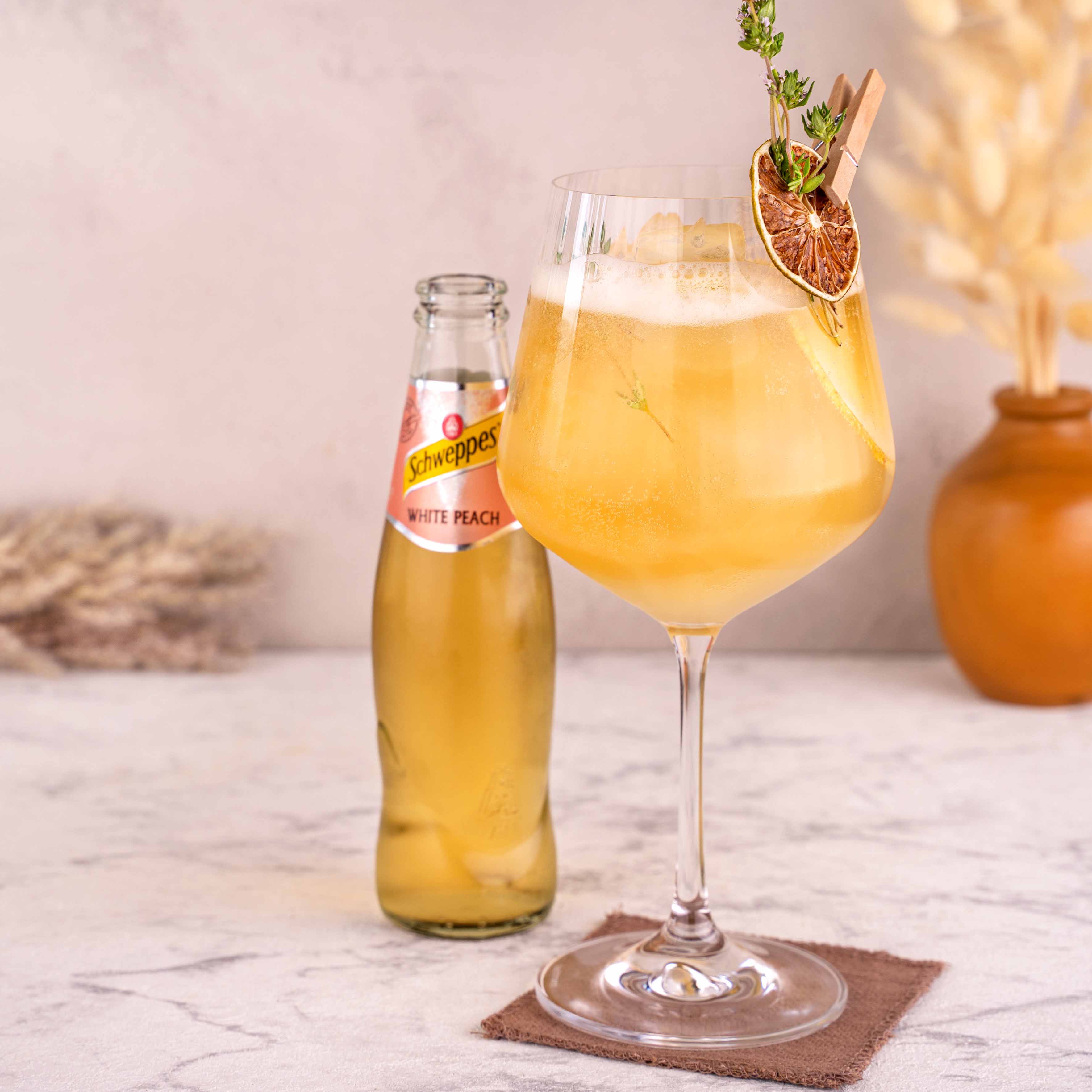 Momotaro Kizaru Spritz Cocktail in großem Glas, dekoriert mit getrockneter Limette, Kräutern und frischer Birne, Schweppes White Peach Flasche daneben, auf hellem Untergrund, Trockenblumen als Dekoration im Hintergrund