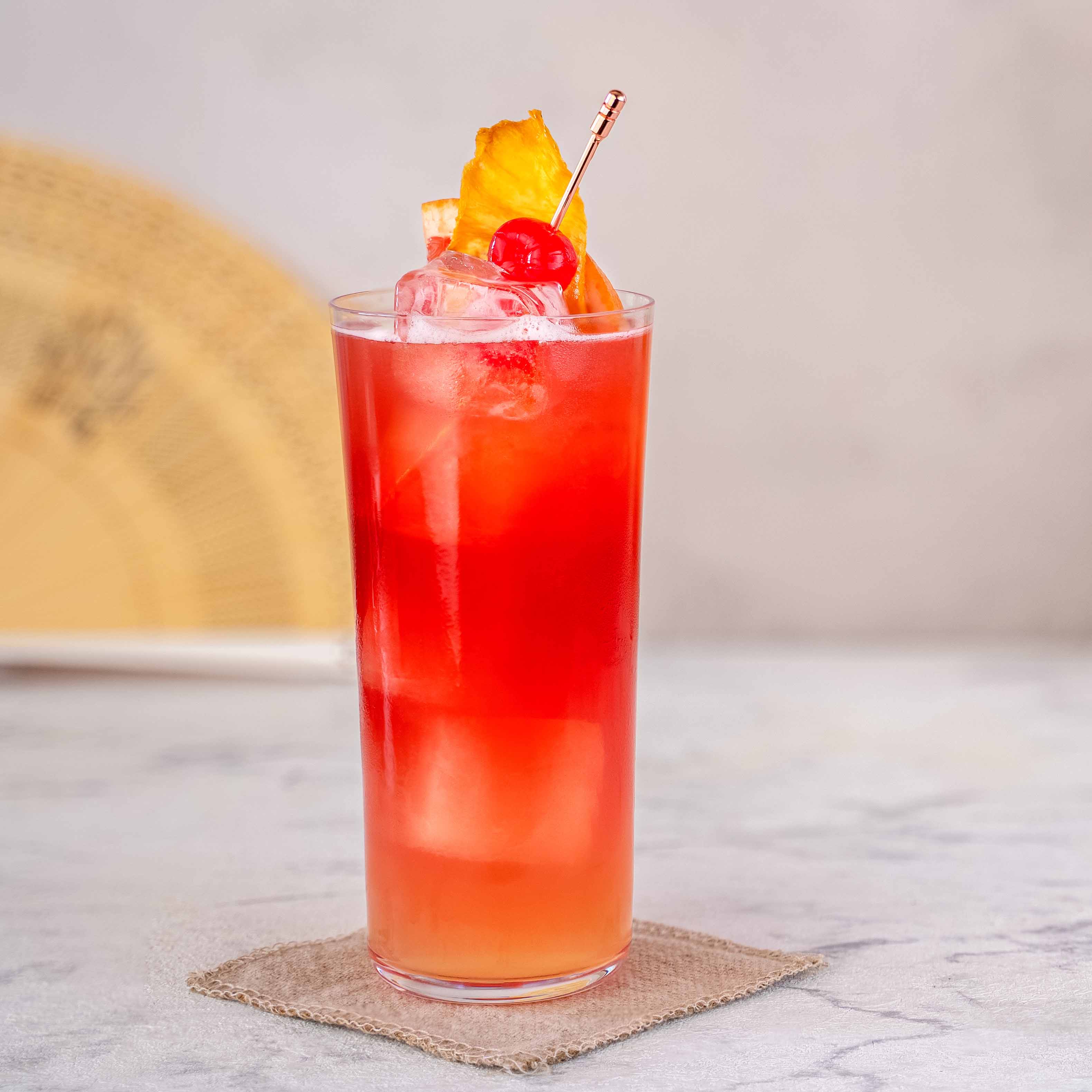 Momotaro Kizaru Tokio Sling Cocktail, roter Cocktail auf Eis, dekoriert mit getrockneter Ananas und Cocktail-Kirsche, auf hellem Untergrund, Japanischer Fächer im Hintergrund