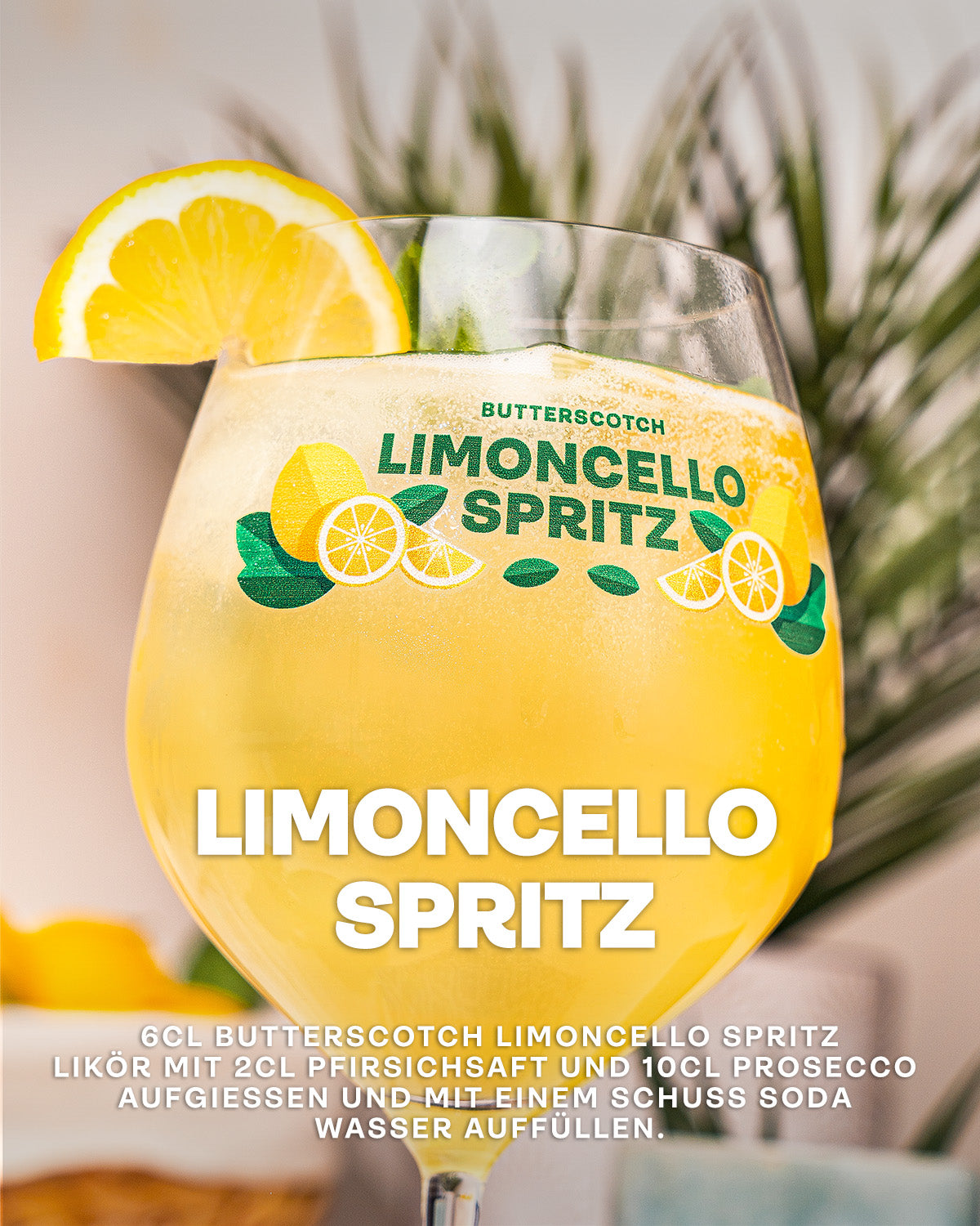 Limoncello Spritz Cocktail, Butterscotch Limoncello Likör mit Pfirsichsaft und Prosecco aufgegossen, Spritz-Glas mit Limoncello Logo im Vordergrund, dekoriert mit frischer Zitrone und Minze, Palmwedel im Hintergrund, auf hellem Untergrund