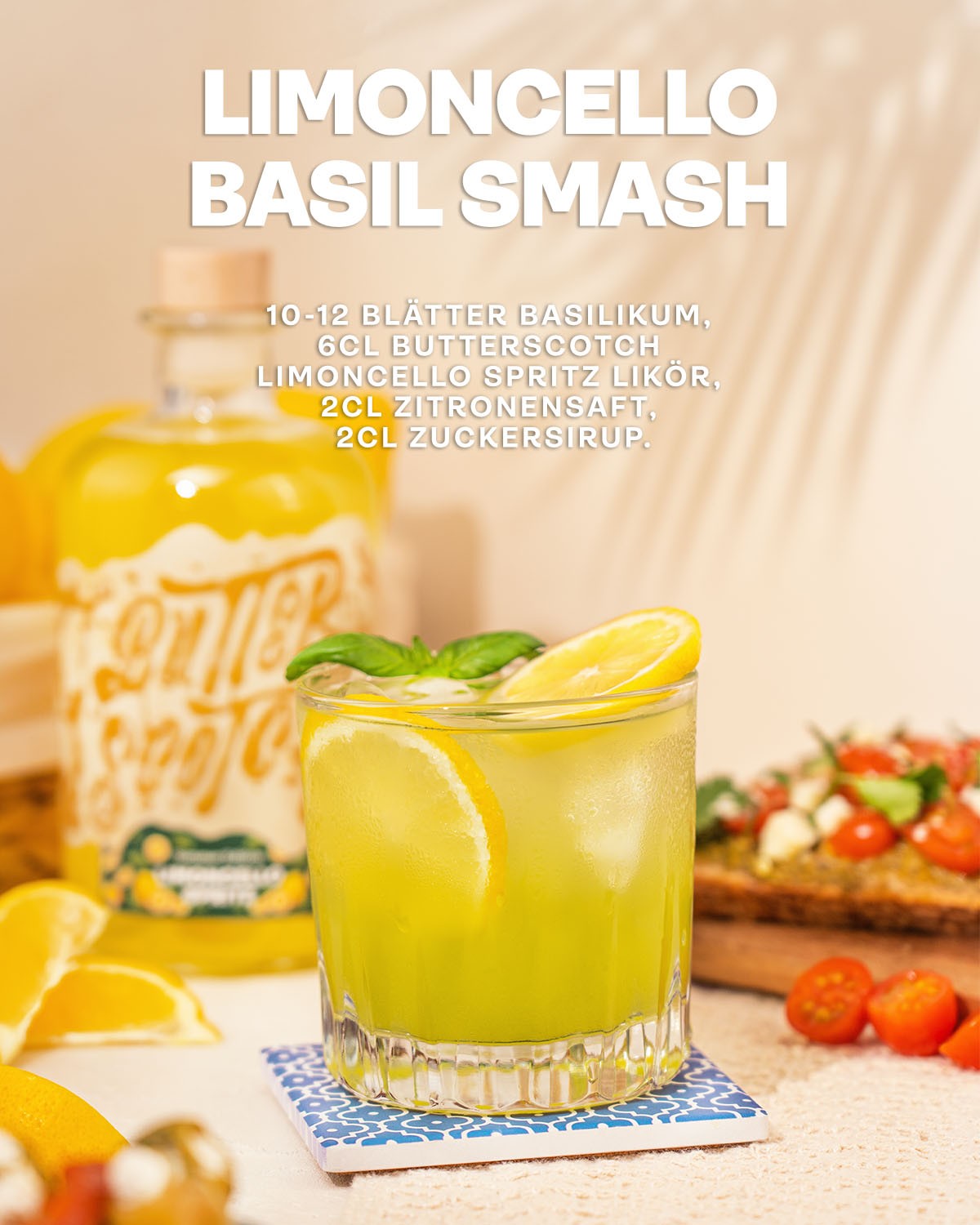 Limoncello Basil Smash, Basilikum, Butterscotch Limoncello Likör, Zitronensaft & Zuckersirup. Cocktail mit Eis, frischen Zitronenscheiben und Minze garniert im Vordergrund zwischen Zitronen und Tomaten, Flasche Limoncello Likör im Hintergrund auf hellem Untergrund