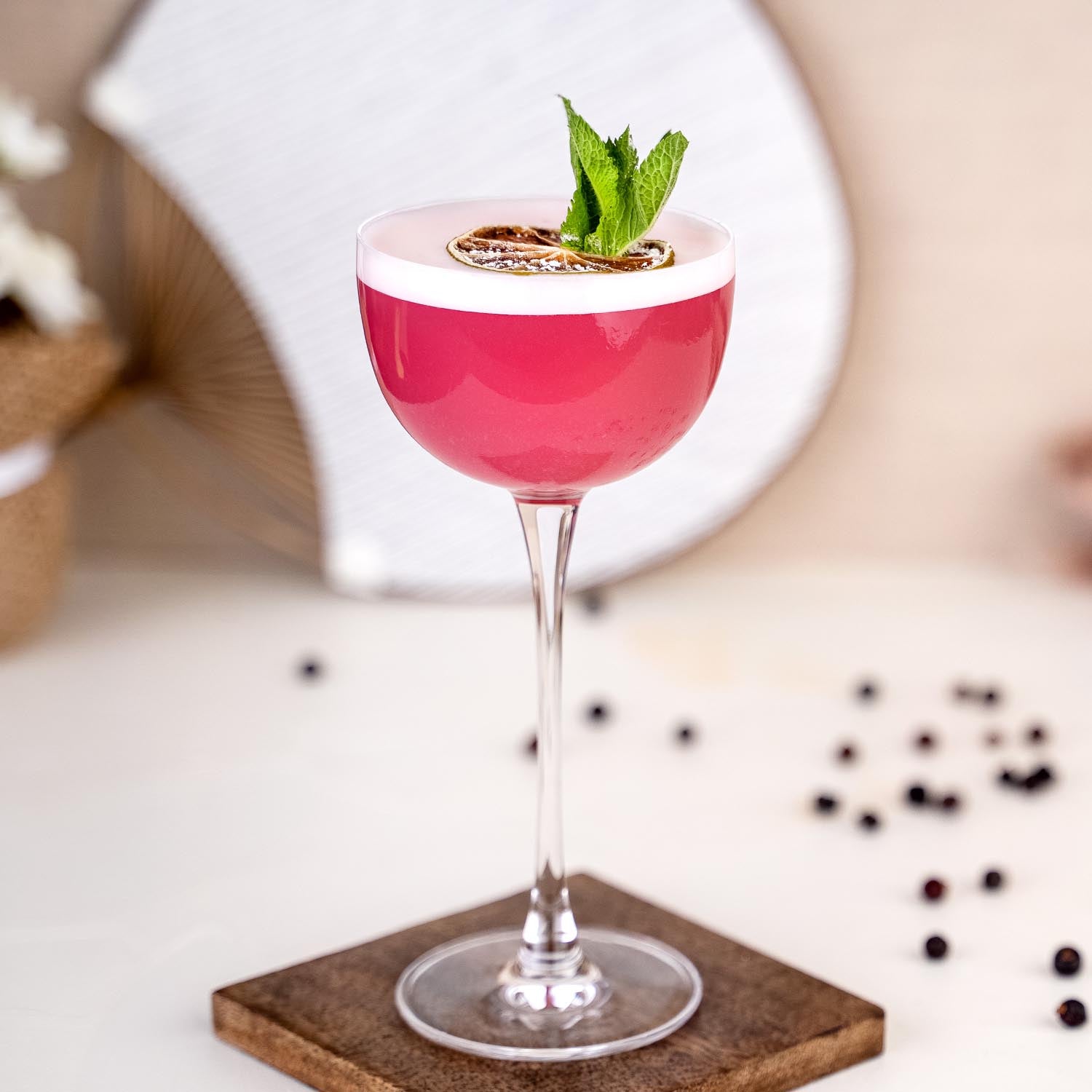 Last Sword Cocktail, pinker Drink in winery Coupetten-Glas mit weißem Schaum, dekoriert mit getrockneter Limette und frischer Minze, vor einem hellen Fächer im Hintergrund