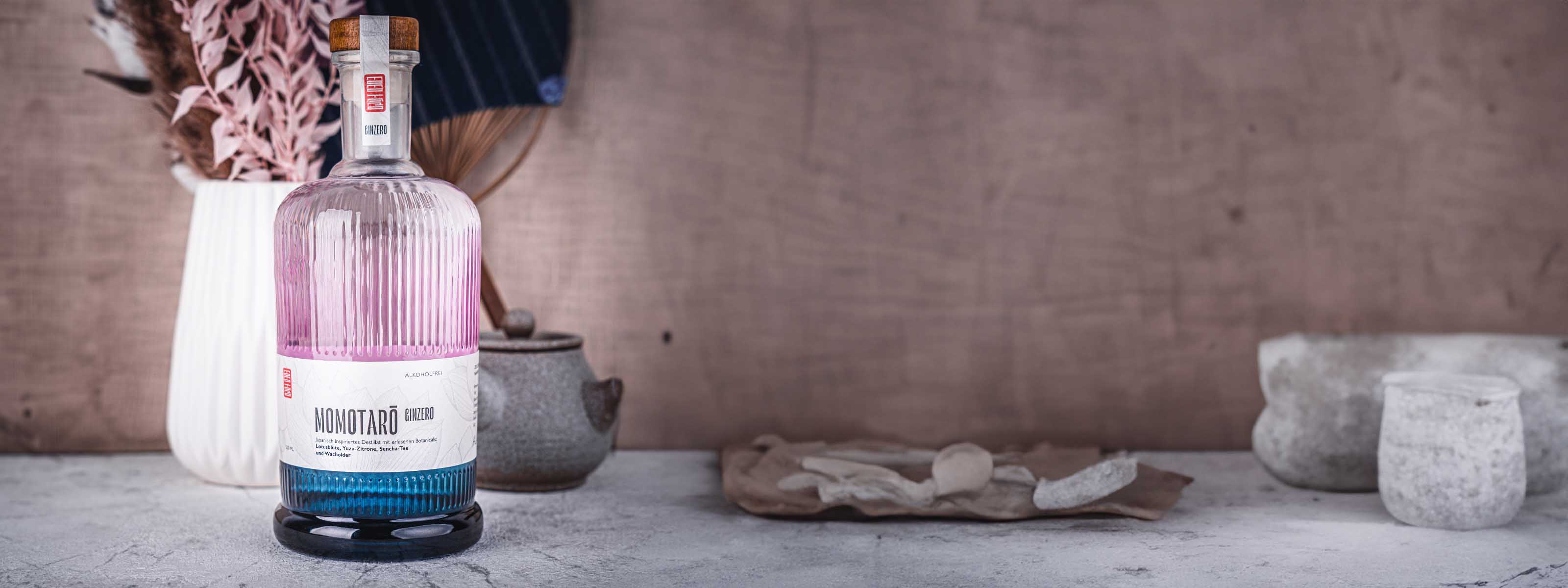 Momotaro Ginzero Flasche links im Bild vor Vase mit Blumen und japanischem Fächer im Hintergrund, auf hellem Untergrund