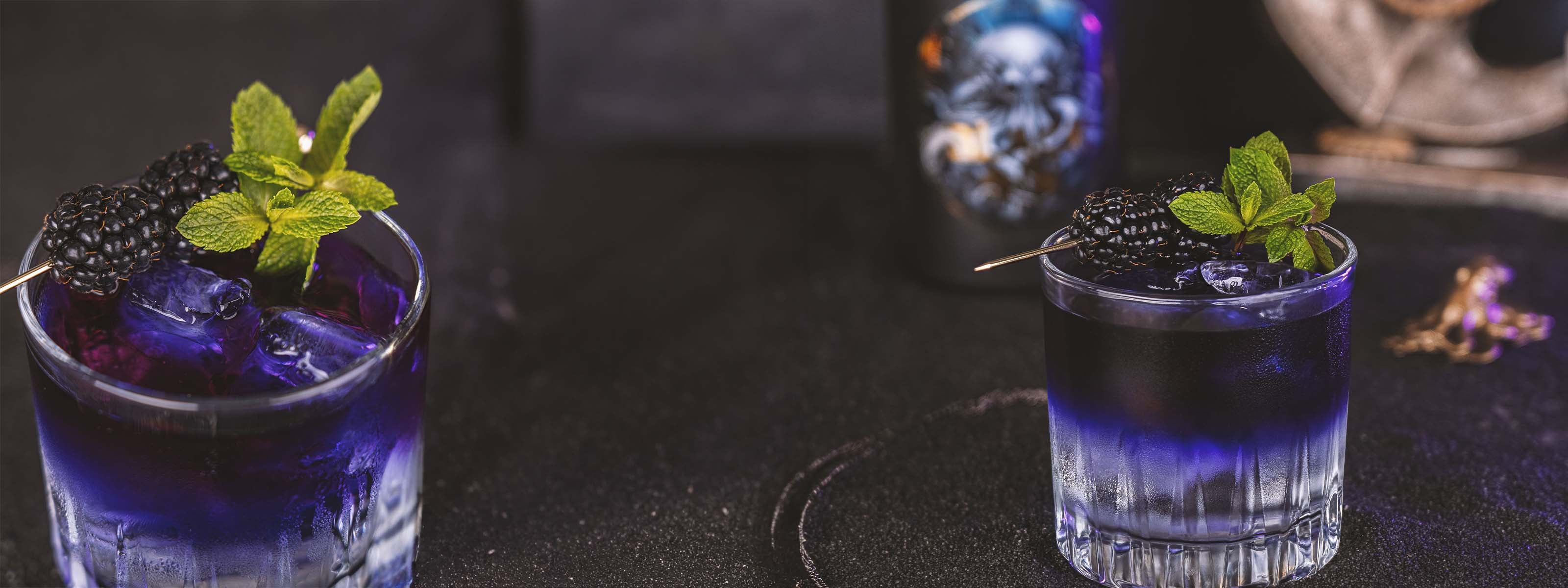 Zwei Floated Gin & Tonic Gläser mit Verlauf vom Tonic Water zum Black gin, vor dunklem Hintergrund mit nautischen Elementen, Drinks garniert mit Minze und Brombeeren