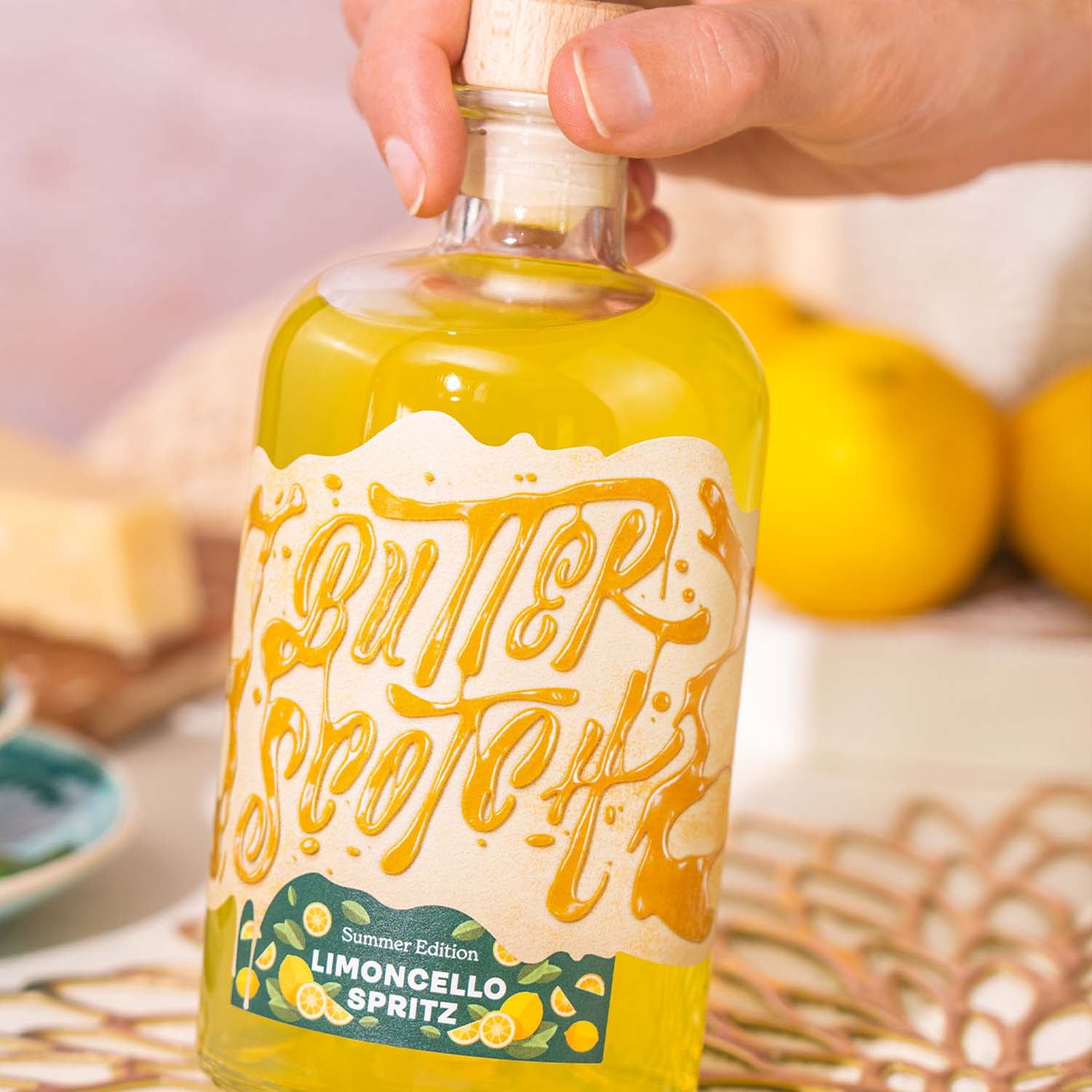 Butterscotch Limoncello Spritz, Flasche mit gelbem Inhalt, vor hellem Hintergrund, frische Zitronen und Picknickteller im Hintergrund, Flasche wird von oben gegriffen