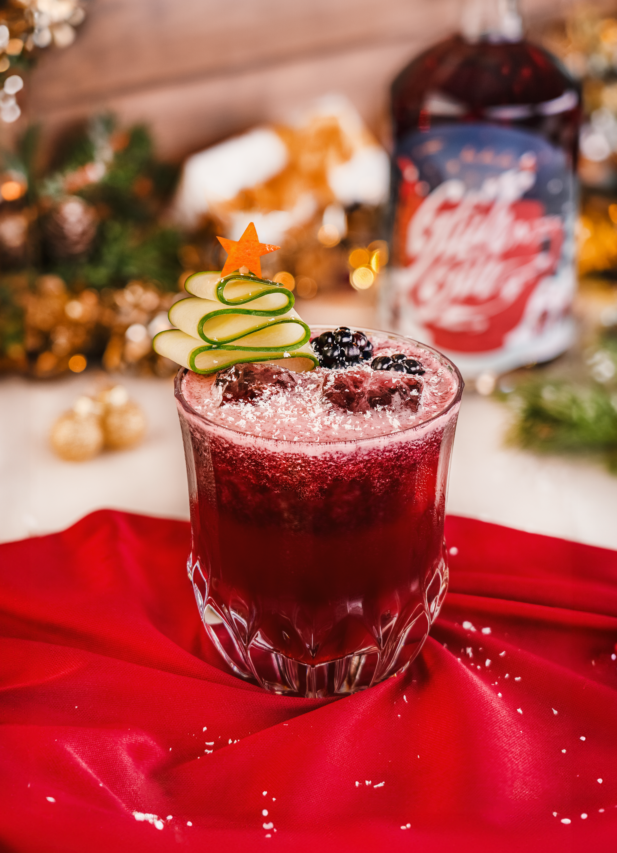 Winter Wonderland Christmas Bramble Cocktail, roter Cocktail in geschliffenem Glas auf rotem Tuch, dekoriert mit frischen Beeren und einem Tannenbaum aus Gurke und Orangen-Stern, lasche & winterliche Dekoration im Hintergrund