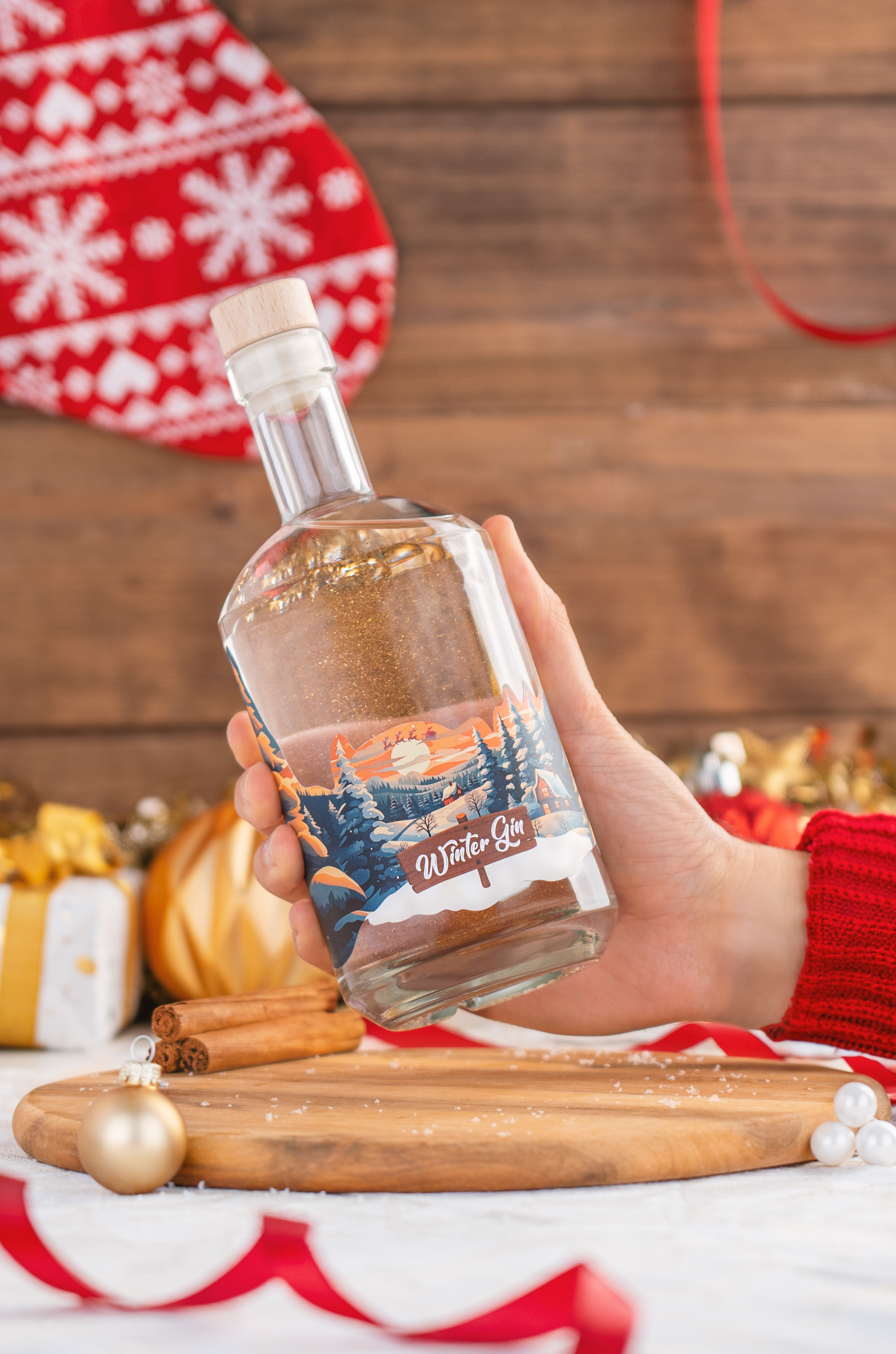 Winter Wonderland Winter Gin mit essbarem Glitzer, Gin mit Zimt, Vanille & Bratapfel, Flasche im Schneekugel-Design, ideal für winterliche Drinks, Flasche im Vordergrund vor Christbaumkugeln und Zimtstangen