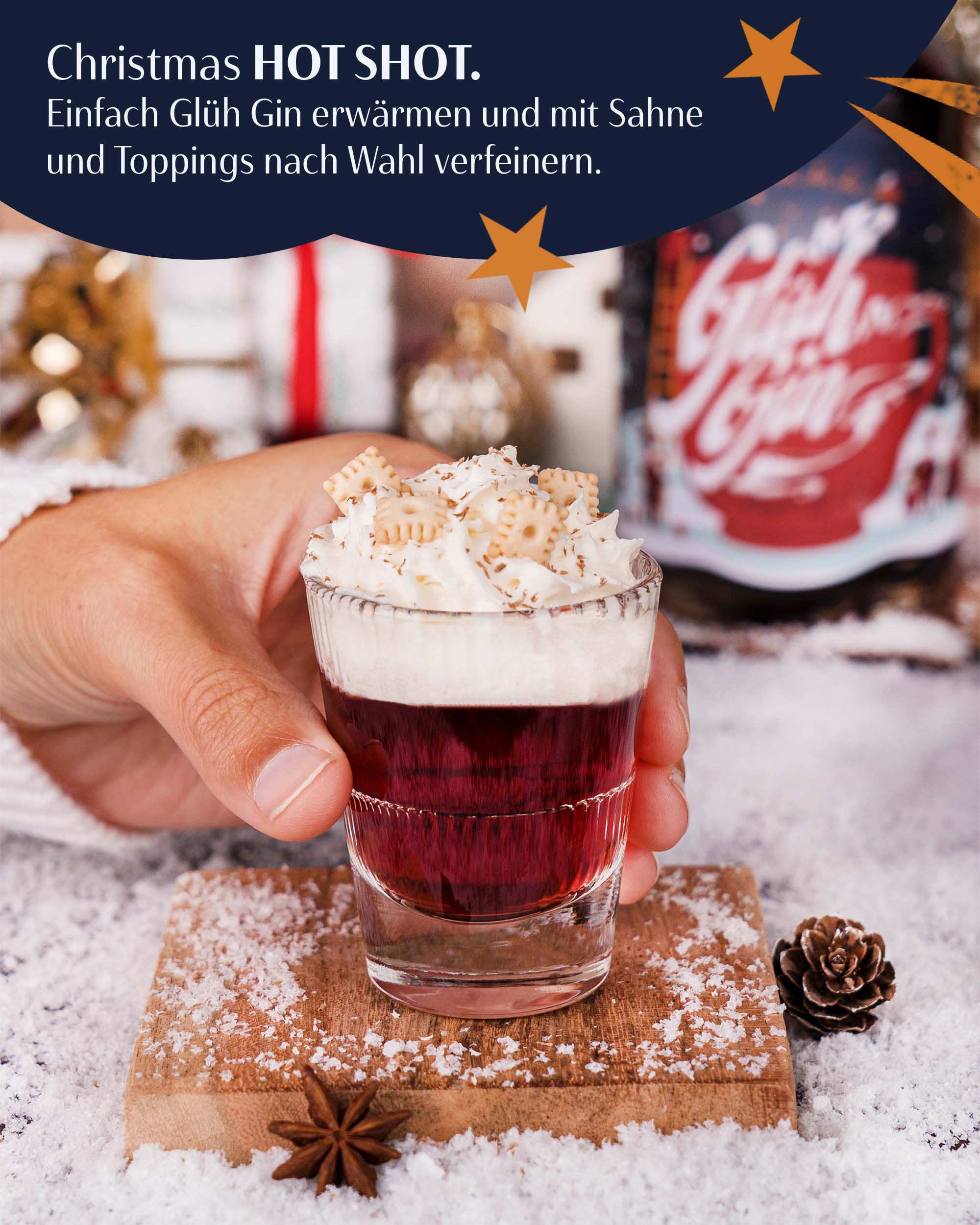 Winter Wonderland Christmas HotShot, Glühgin im Shotglas, dekoriert mit Sahnehaube und Keksen, wird von einer Hand gegriffen, auf Holzbrett im Kunstschnee, Flasche Glühgin unscharf im Hintergrund