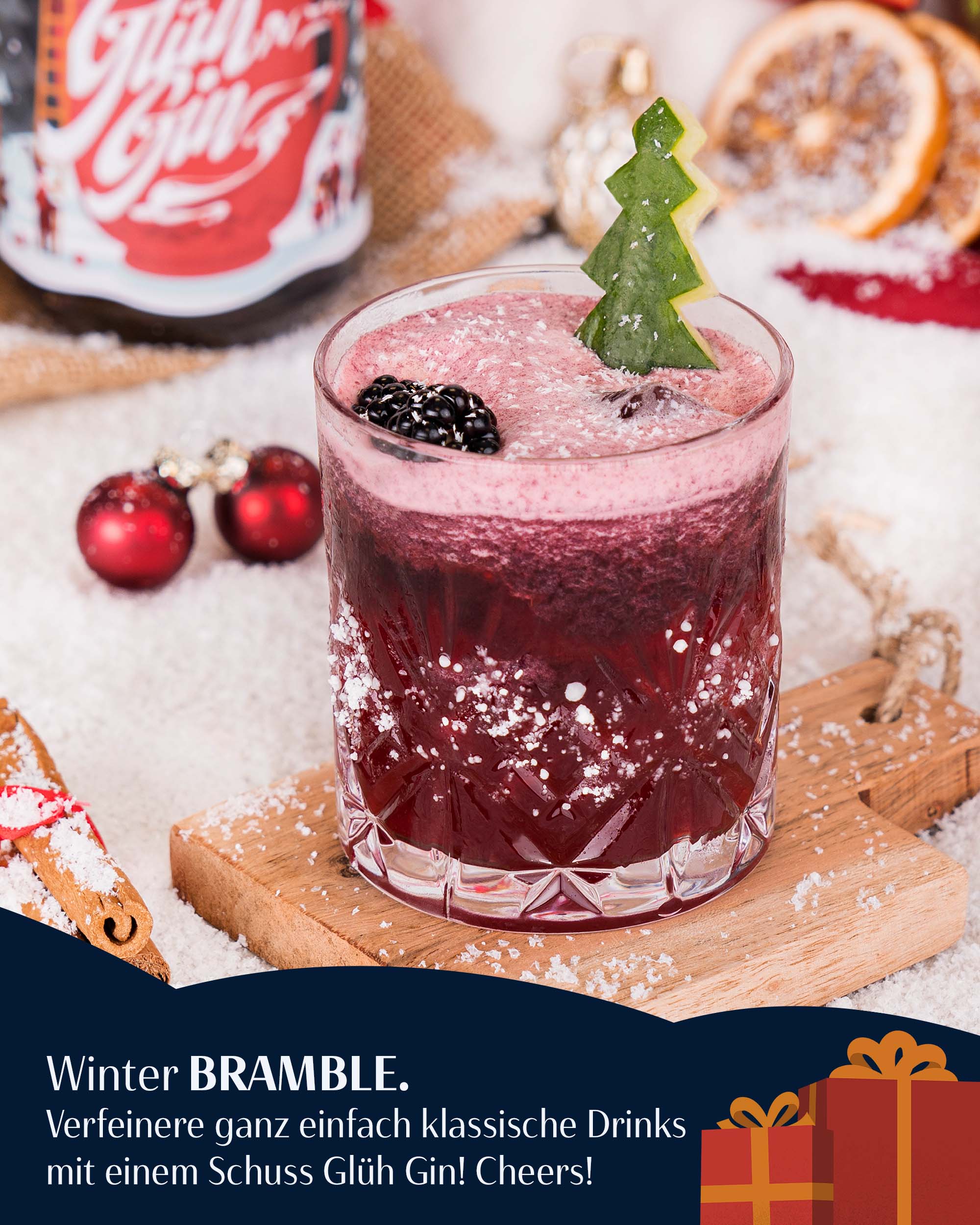 Winter Wonderland Bramble Cocktail, dunkelroter Cocktail mit Schaumkrone, dekoriert mit Gurke in Tannenbaum-Form geschnitzt und frischen Brombeeren, auf Holzbrett in Kunstschnee, Flasche Glüh Gin unscharf im Hintergrund